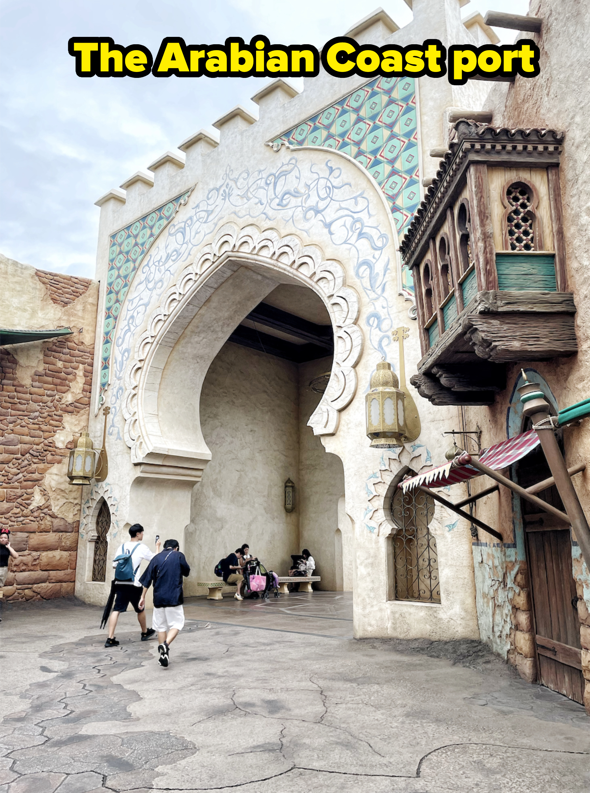 People walking near an ornate arched entrance with intricate designs in a theme park; a group is sitting inside the arch
