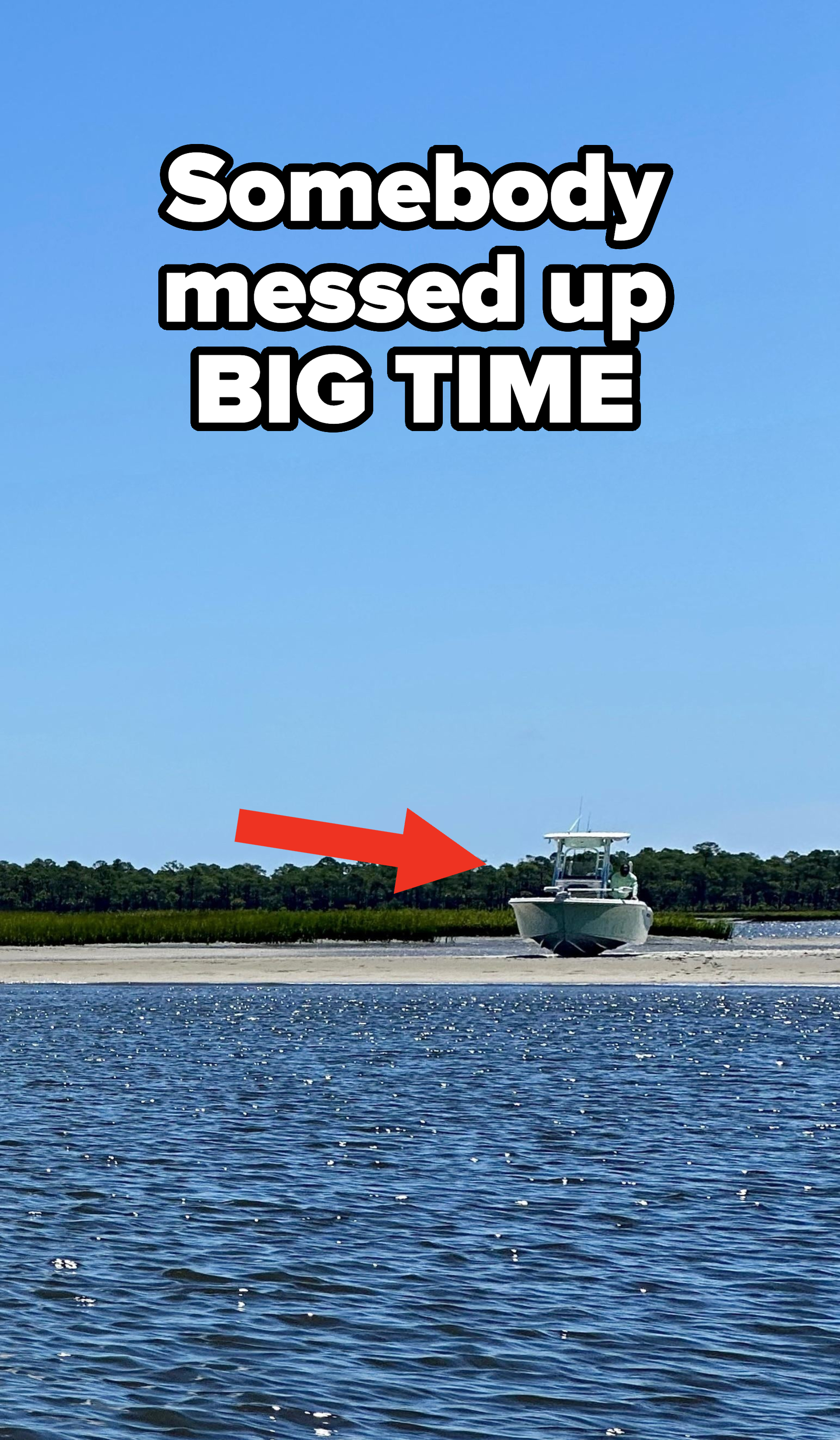 A boat is grounded on a sandbar near the shore with a backdrop of trees and water. The sky is clear and blue