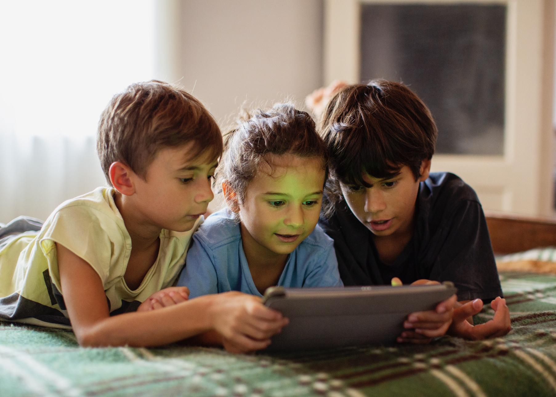 Three children are lying on a bed, sharing and focusing on a tablet screen together