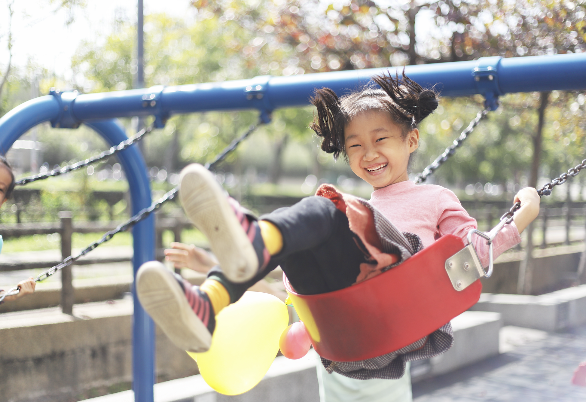 A young girl laughing and joyfully swinging on a playground swing, holding a balloon. Trees and park benches can be seen in the background