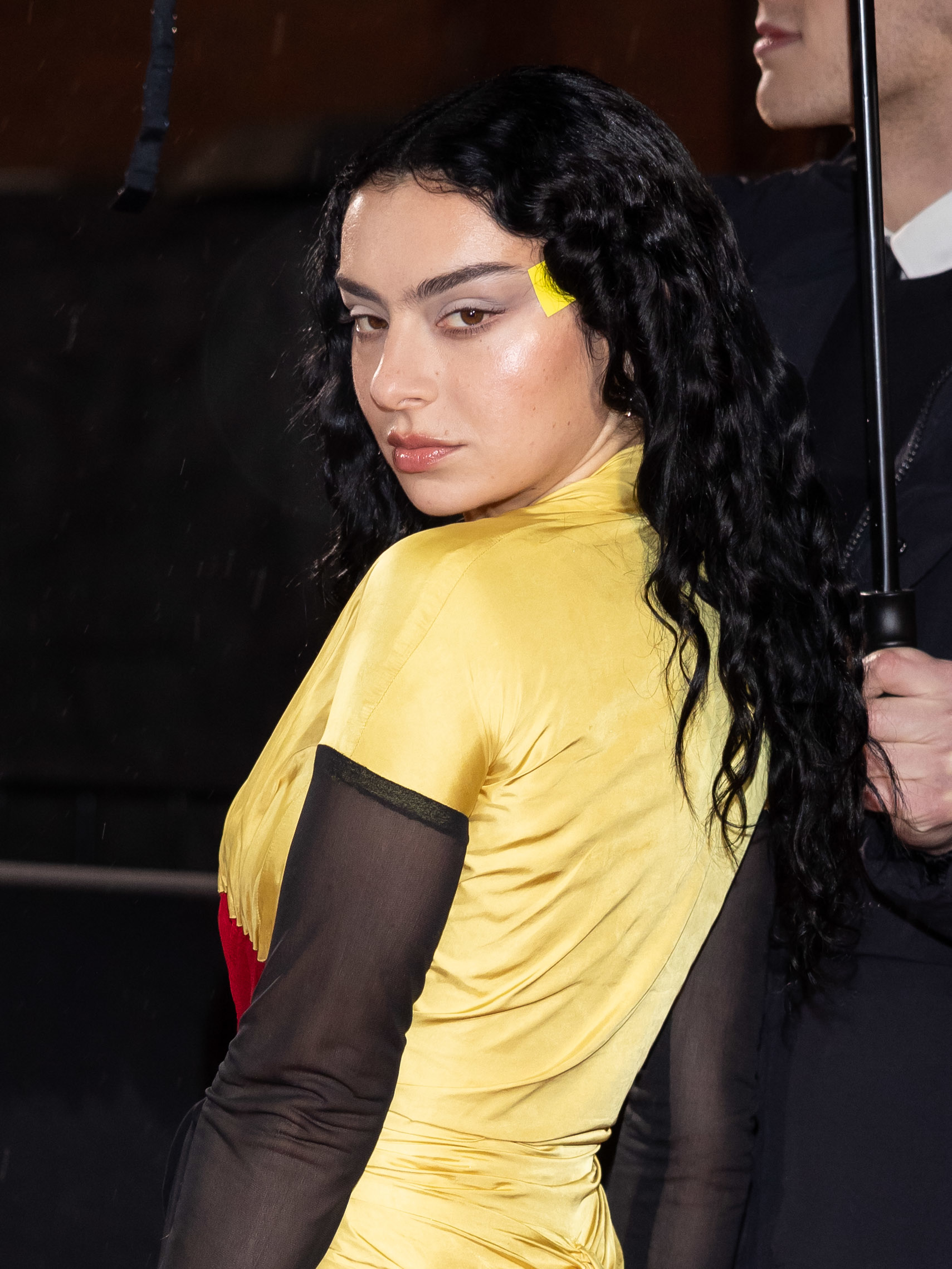 Charli XCX, wearing a unique yellow and black outfit with a red accent, poses for a photo at an event