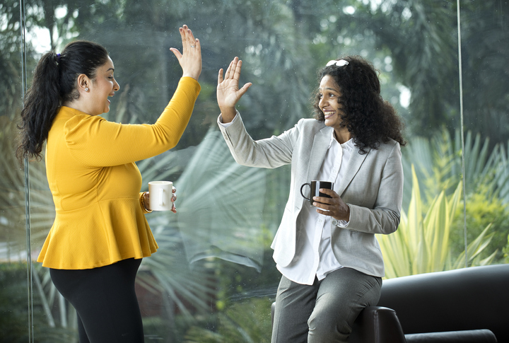 Two women, one in a yellow top and the other in a light-colored suit, are smiling and giving each other a high-five while holding coffee mugs