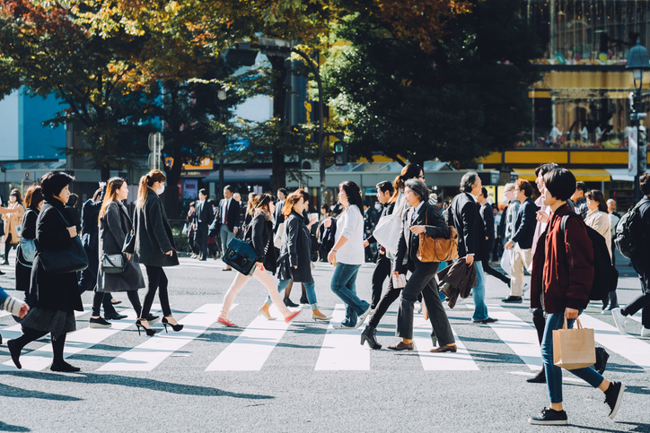 People crossing a busy street at a crosswalk in an urban area, with some carrying shopping bags and others in office attire, captured during daytime