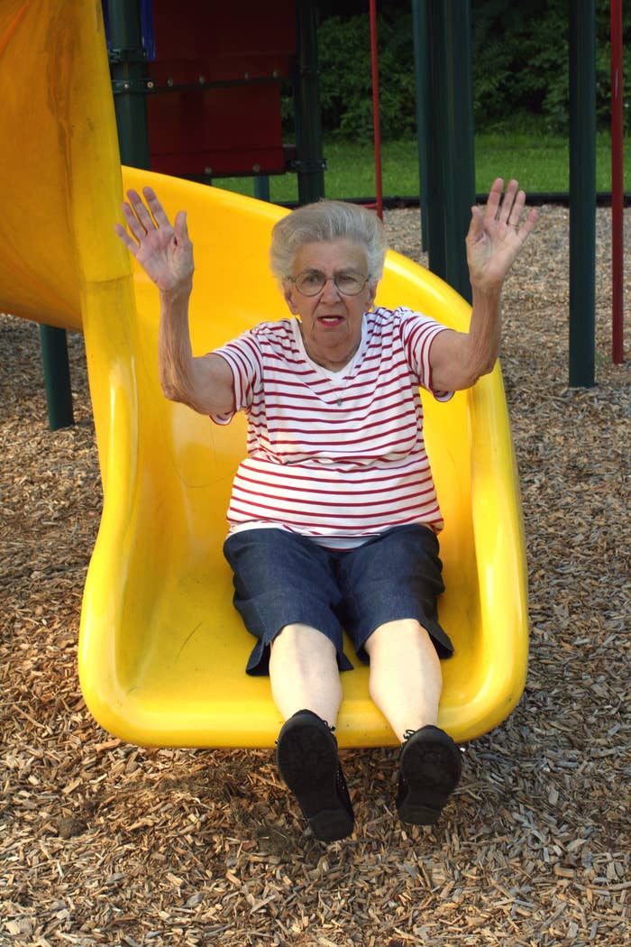 An elderly woman in a striped shirt and denim shorts slides down a yellow playground slide with her hands raised in excitement