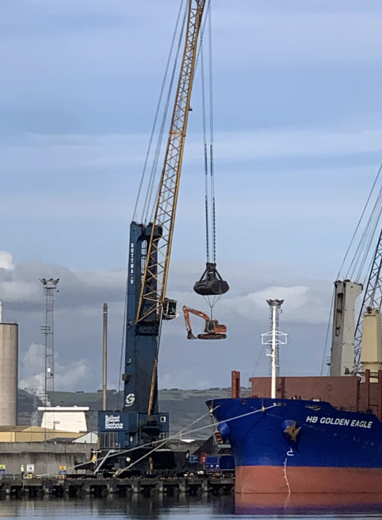 A large crane lifts an excavator onto a blue cargo ship named &quot;HB Golden Eagle&quot; at a harbor. Industrial facilities and other machinery are in the background