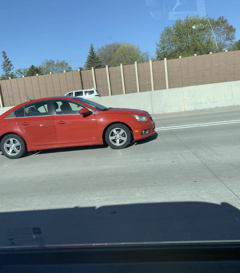 A red car is driving on a highway beside a concrete barrier. Trees and another car are visible in the background