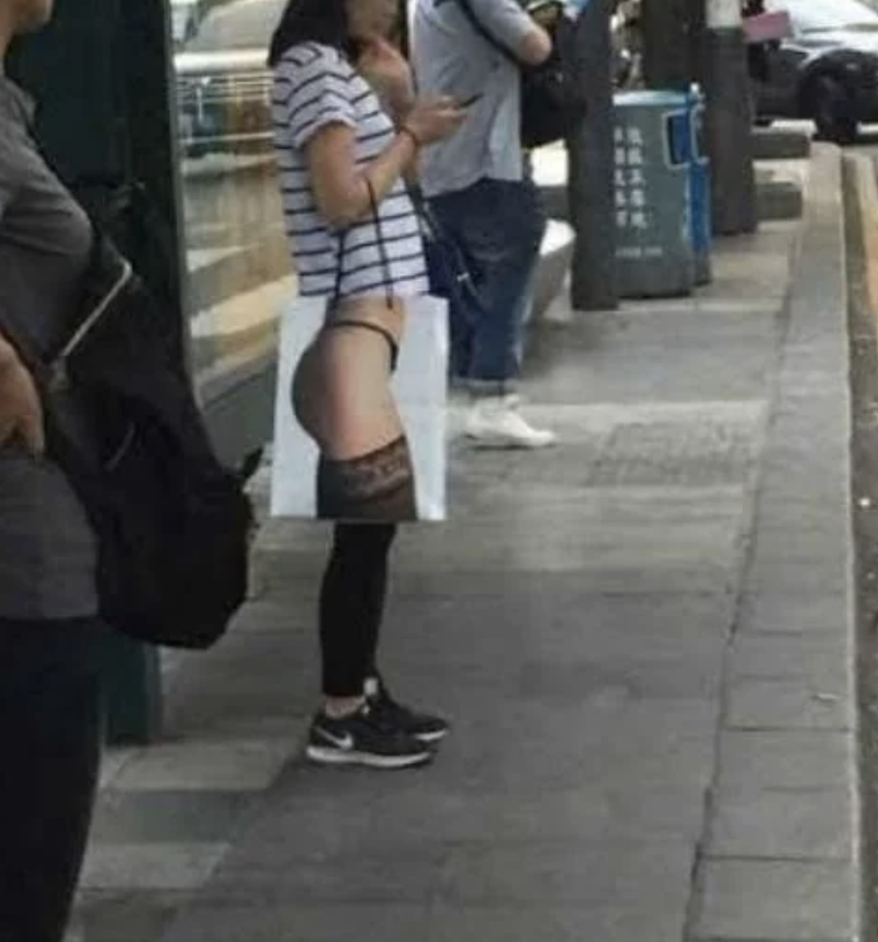 Woman wearing a striped shirt stands at a bus stop with a humorous tote bag, featuring an image of legs in stockings aligned with her lower body