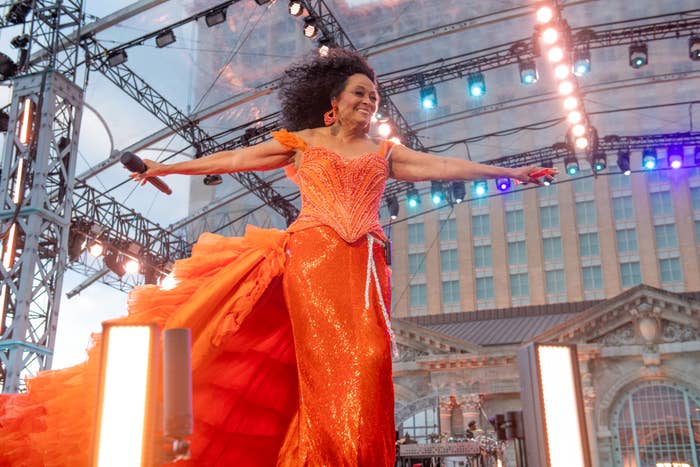 Diana Ross performs on stage wearing a sparkling sequined gown with a ruffled train, arms outstretched. The backdrop includes a building and event lighting