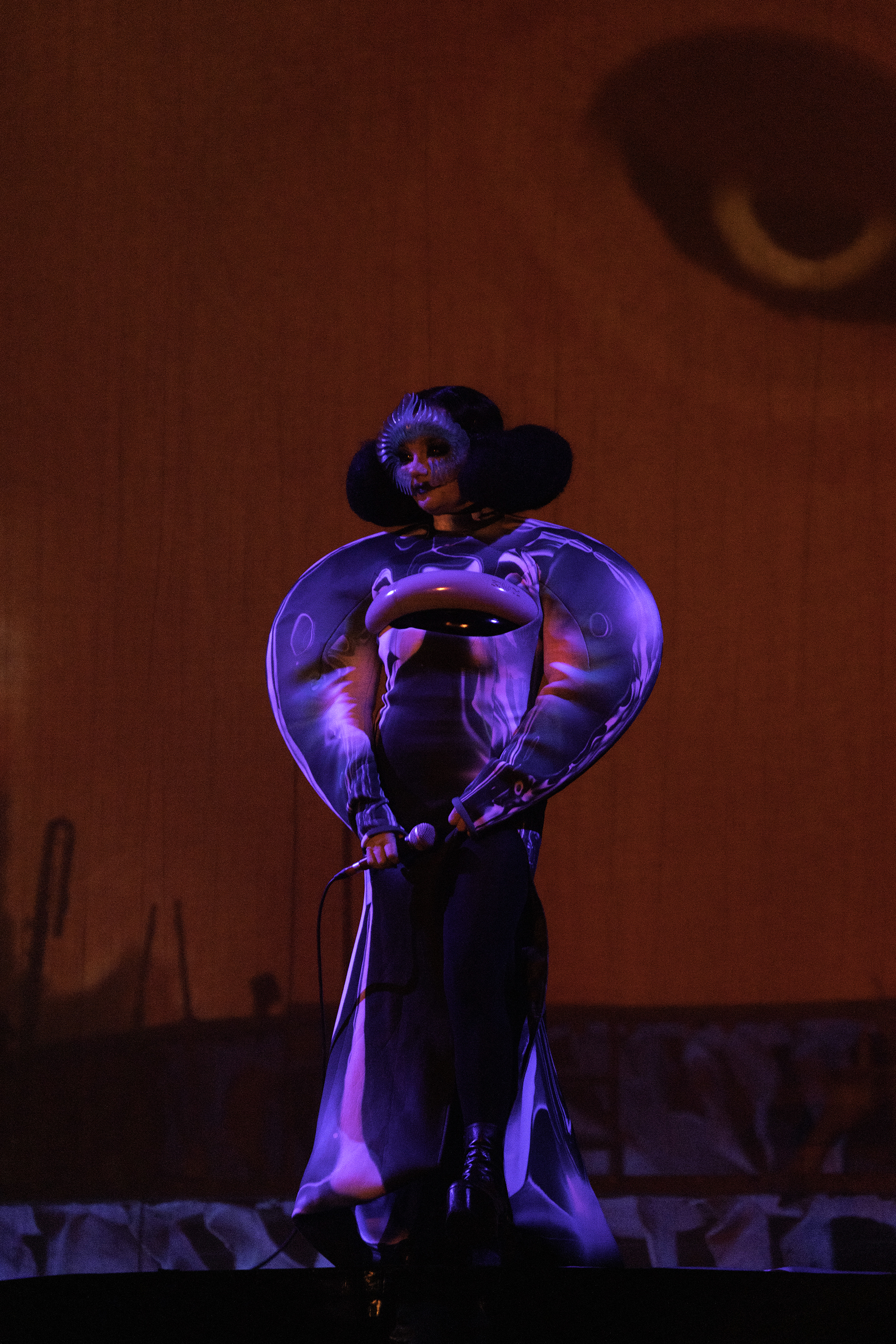 Bjork performs on stage wearing avant-garde costume with large sculptural shoulders and headpiece, in front of a large eye backdrop