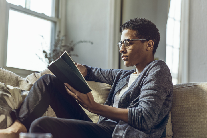 A person with glasses is sitting on a couch and reading a book. The living space includes a window in the background