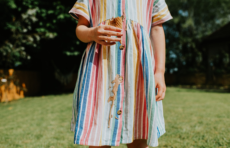 A young person standing on grass, wearing a striped dress with a large stain on the front. The person&#x27;s face is not visible