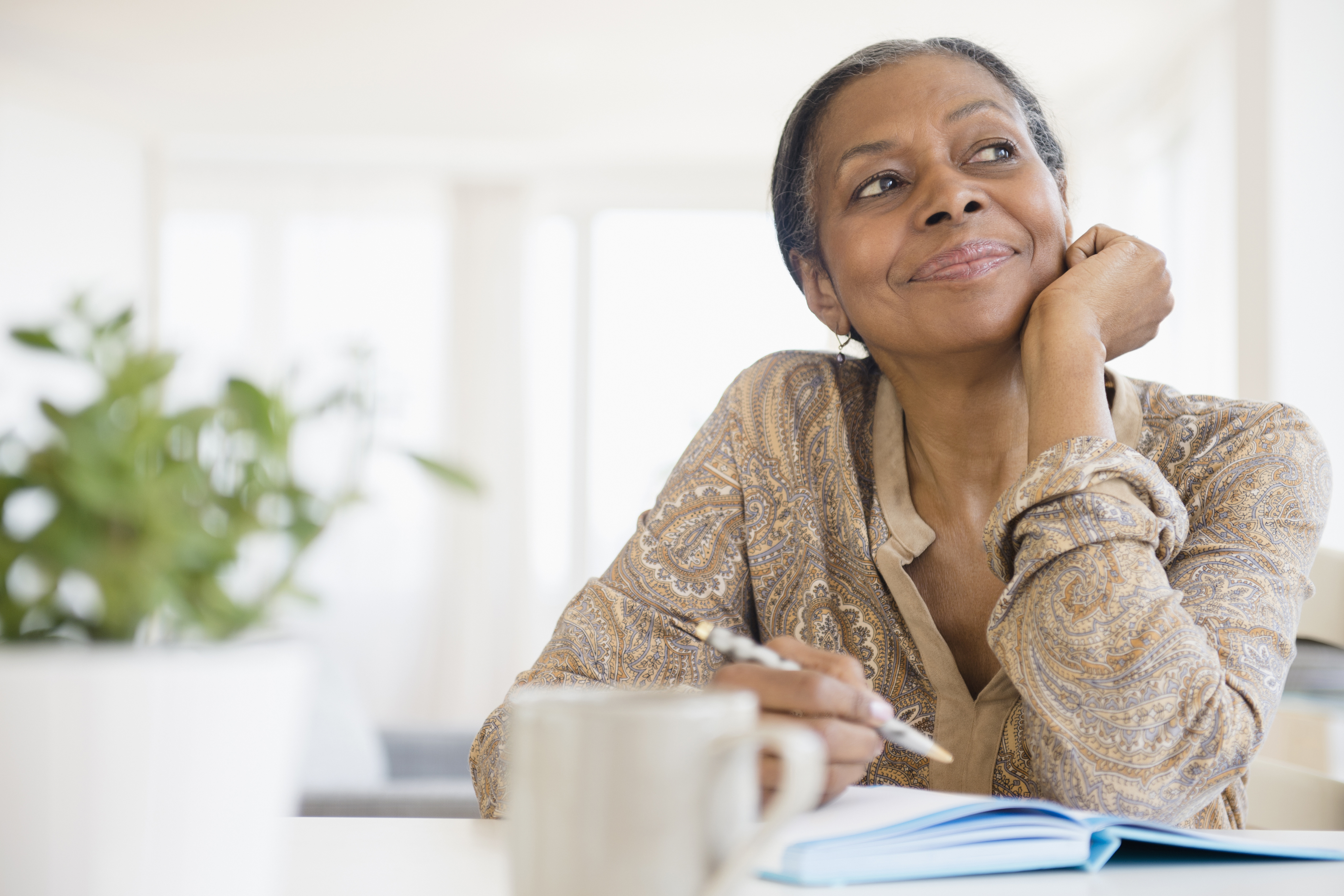 A person sits at a desk, smiling thoughtfully with a pen in hand, beside an open notebook and a plant in the foreground