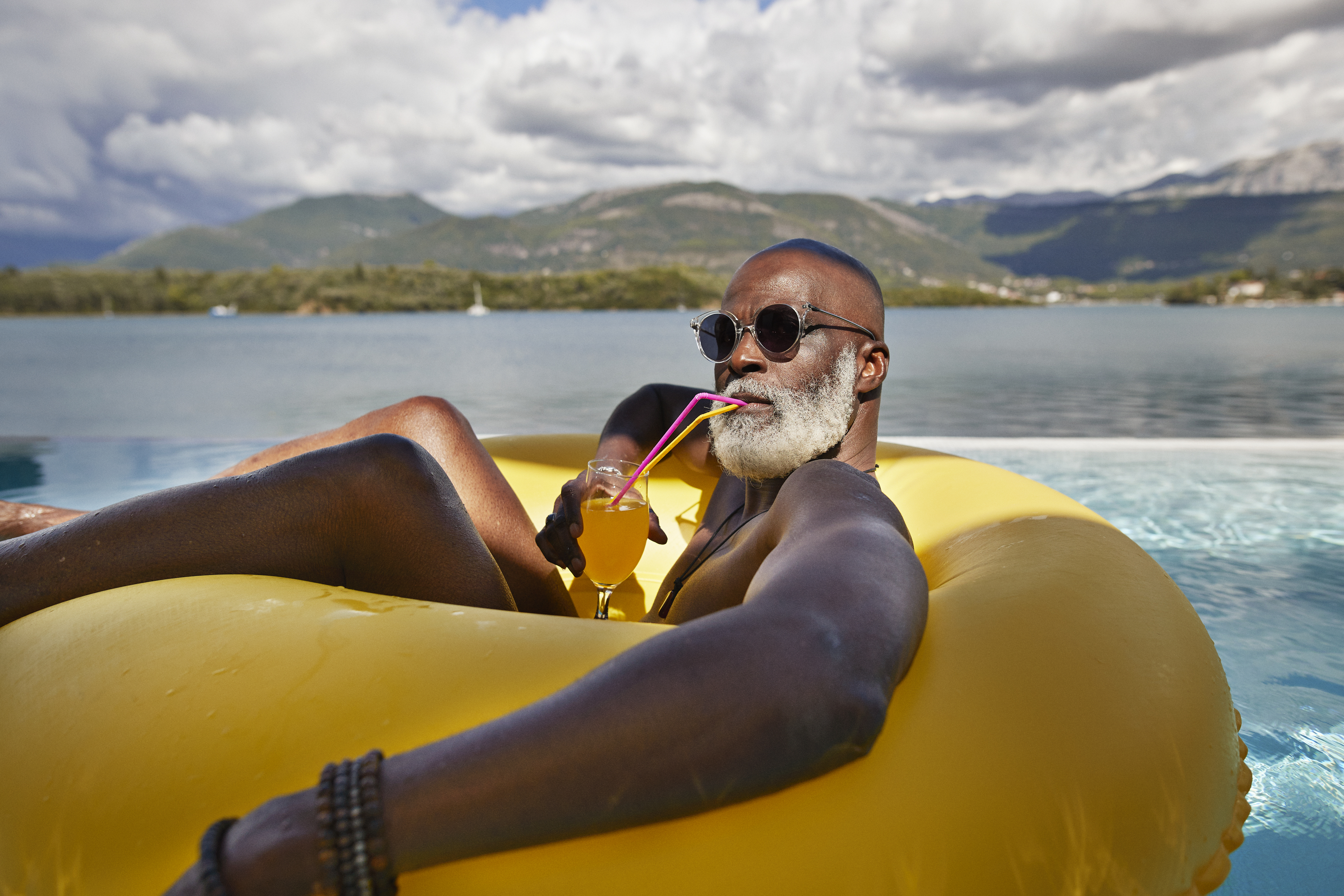 Man with white beard and sunglasses, relaxing on a yellow pool float, sipping a drink with a straw, with mountains and lake in the background