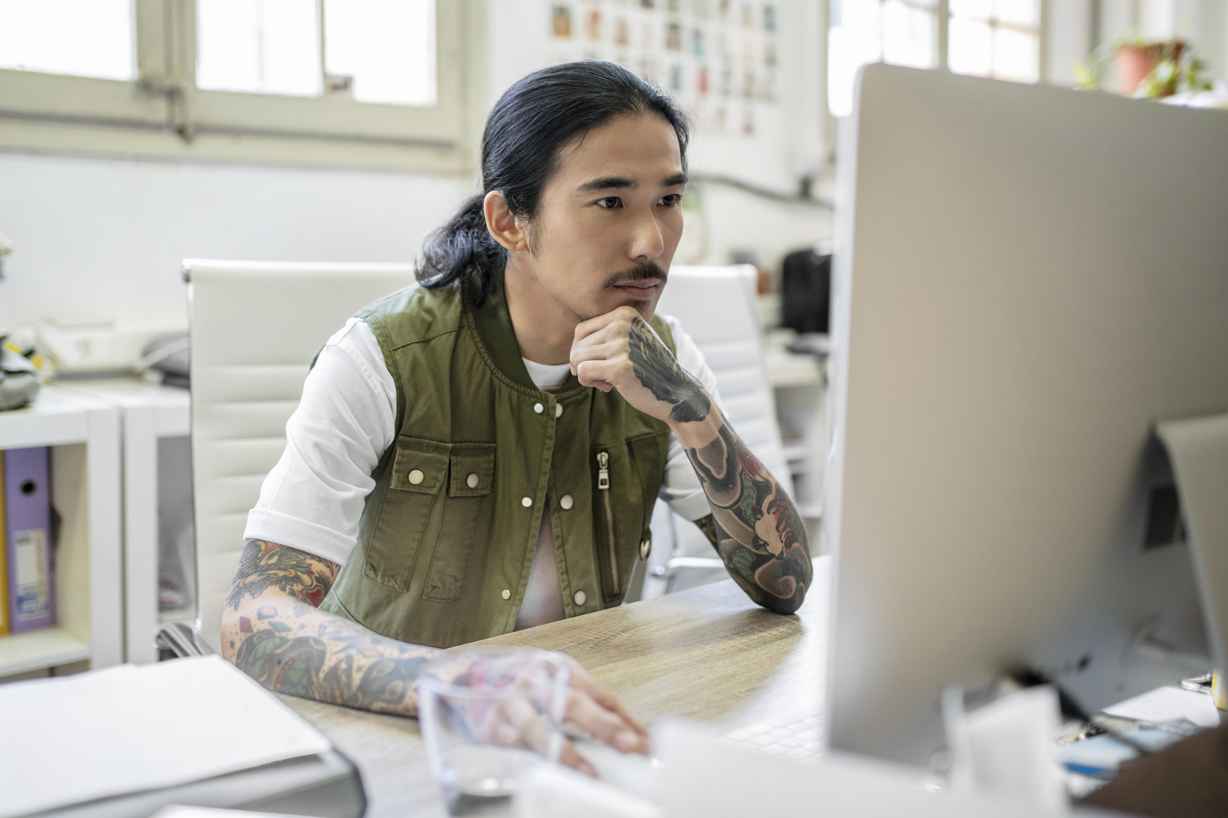 A person with long hair and tattoos works at a desk, intently looking at a large computer monitor. Office supplies and a notebook are on the desk