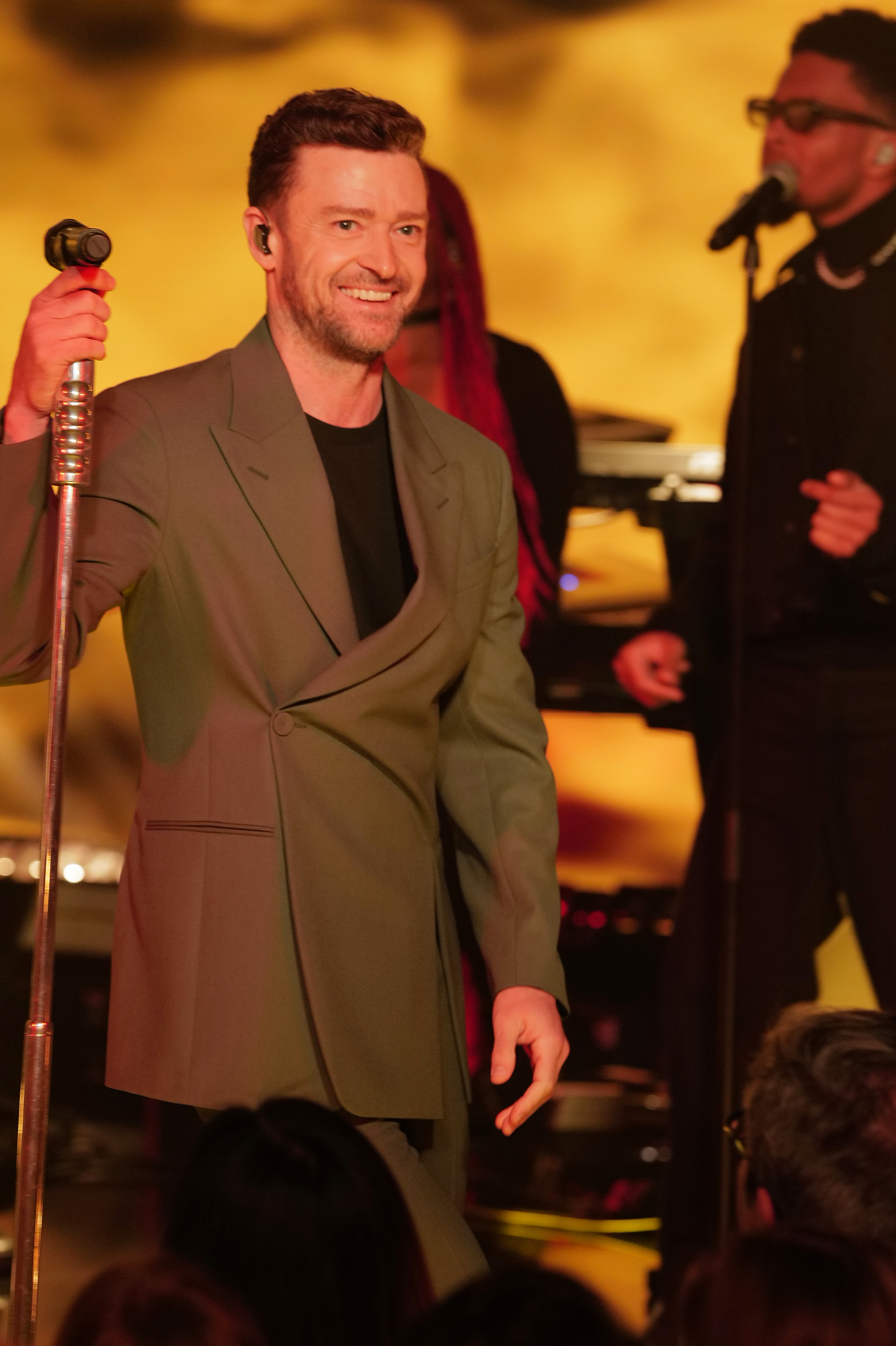 Justin Timberlake performs on stage wearing a green suit, holding a microphone. Background performers are visible in the scene