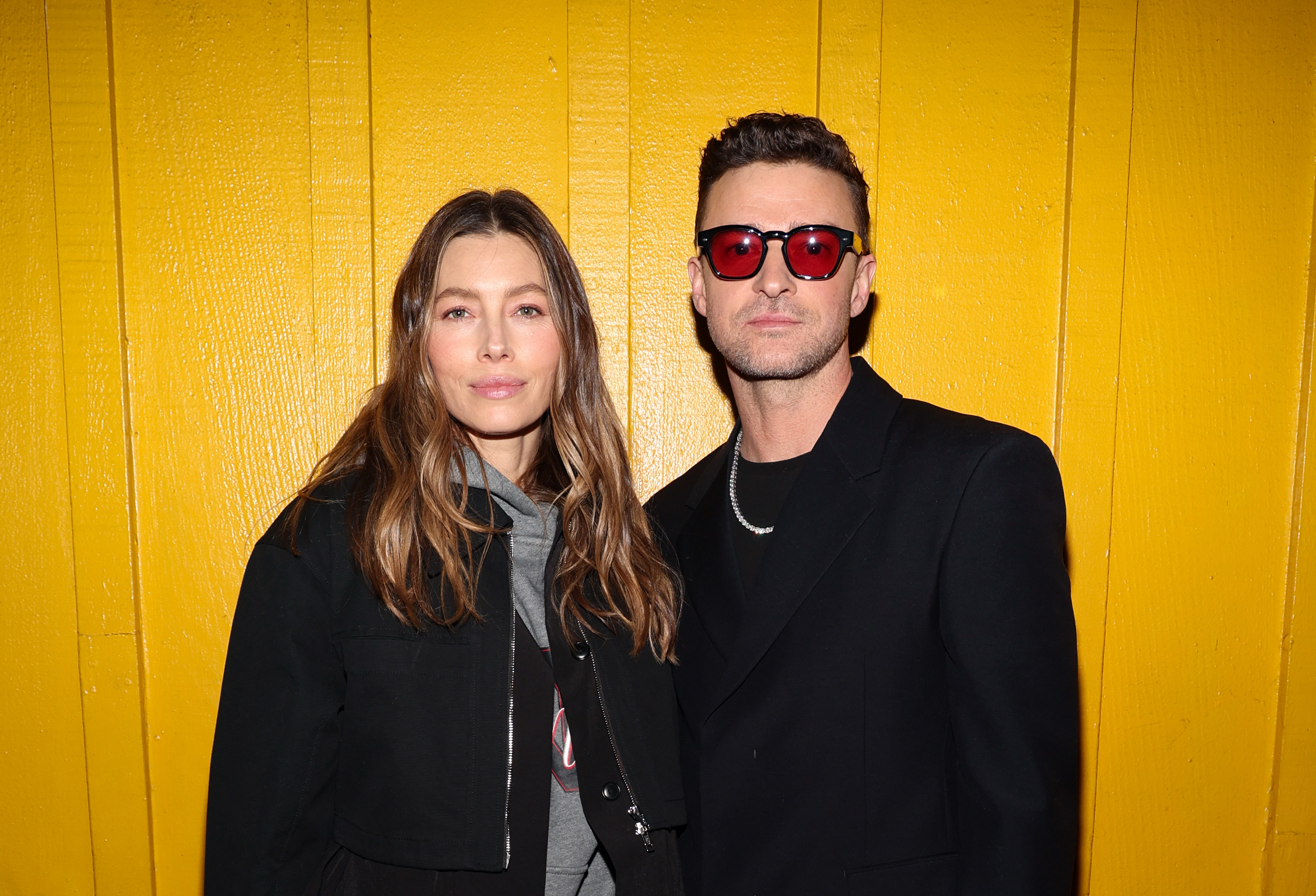Jessica Biel, in casual wear, and Justin Timberlake, in a trendy black jacket and red sunglasses, pose together against a yellow background