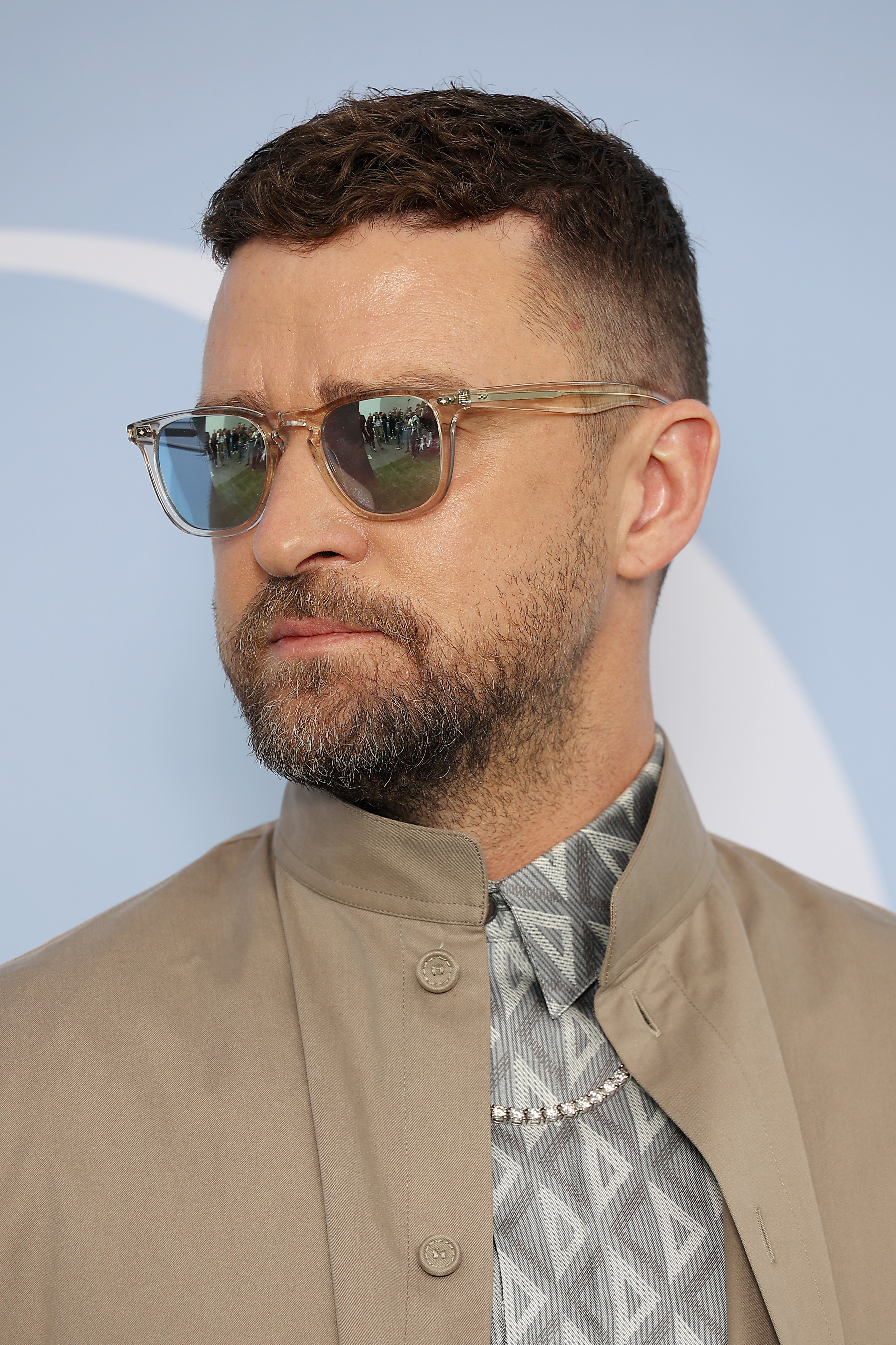 Justin Timberlake wearing a patterned shirt with a high neckline and a beige jacket, accessorized with sunglasses and a necklace