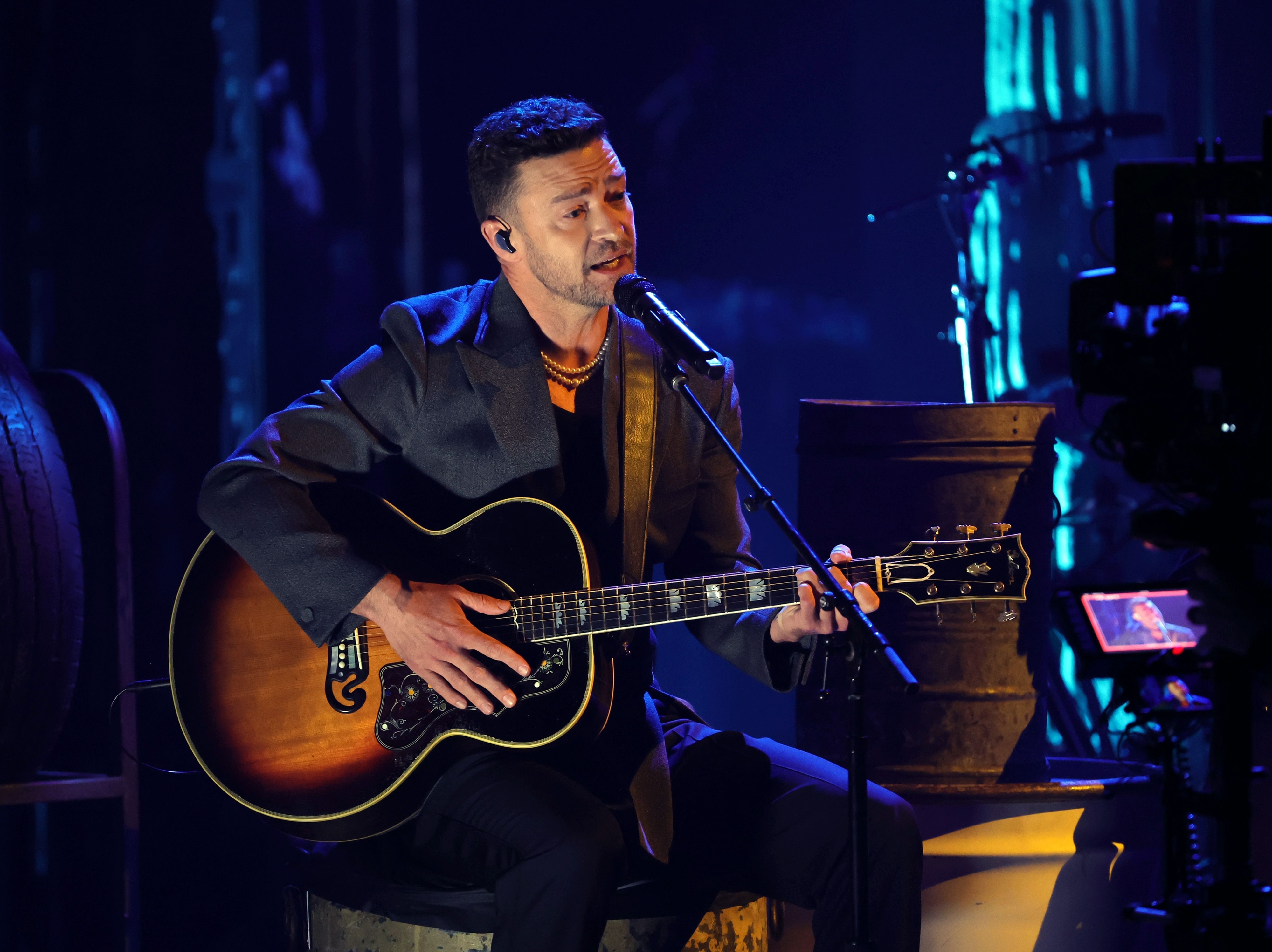 Justin Timberlake performs on stage with an acoustic guitar and a microphone, dressed in a stylish suit jacket