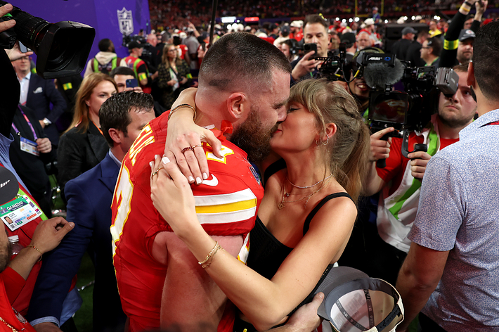Travis Kelce, in a football uniform, and Taylor Swift, in a black dress, embrace and kiss amidst a crowd at a sports event