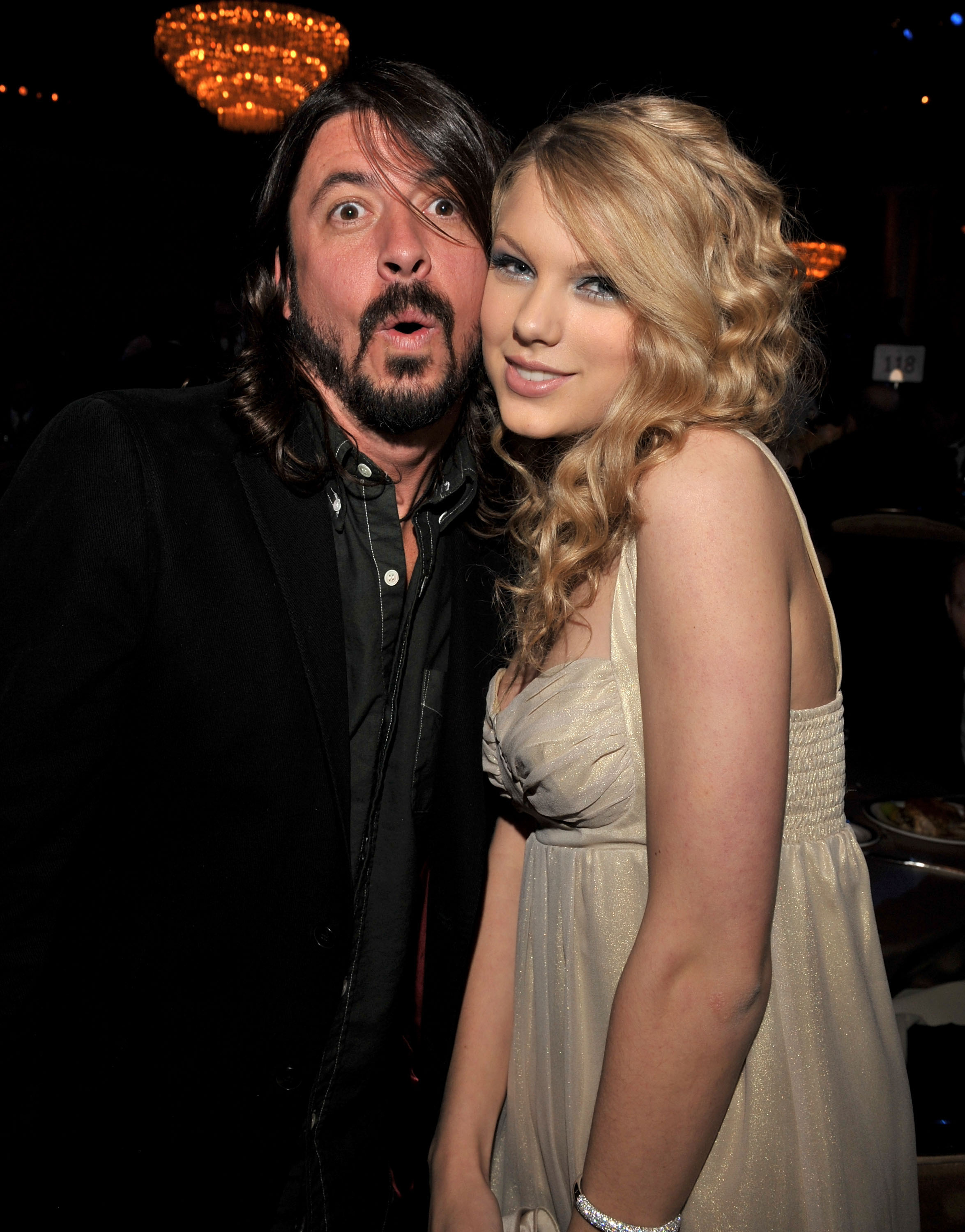 Taylor Swift in a sleeveless dress posing with Dave Grohl in a suit at an event. Both are smiling