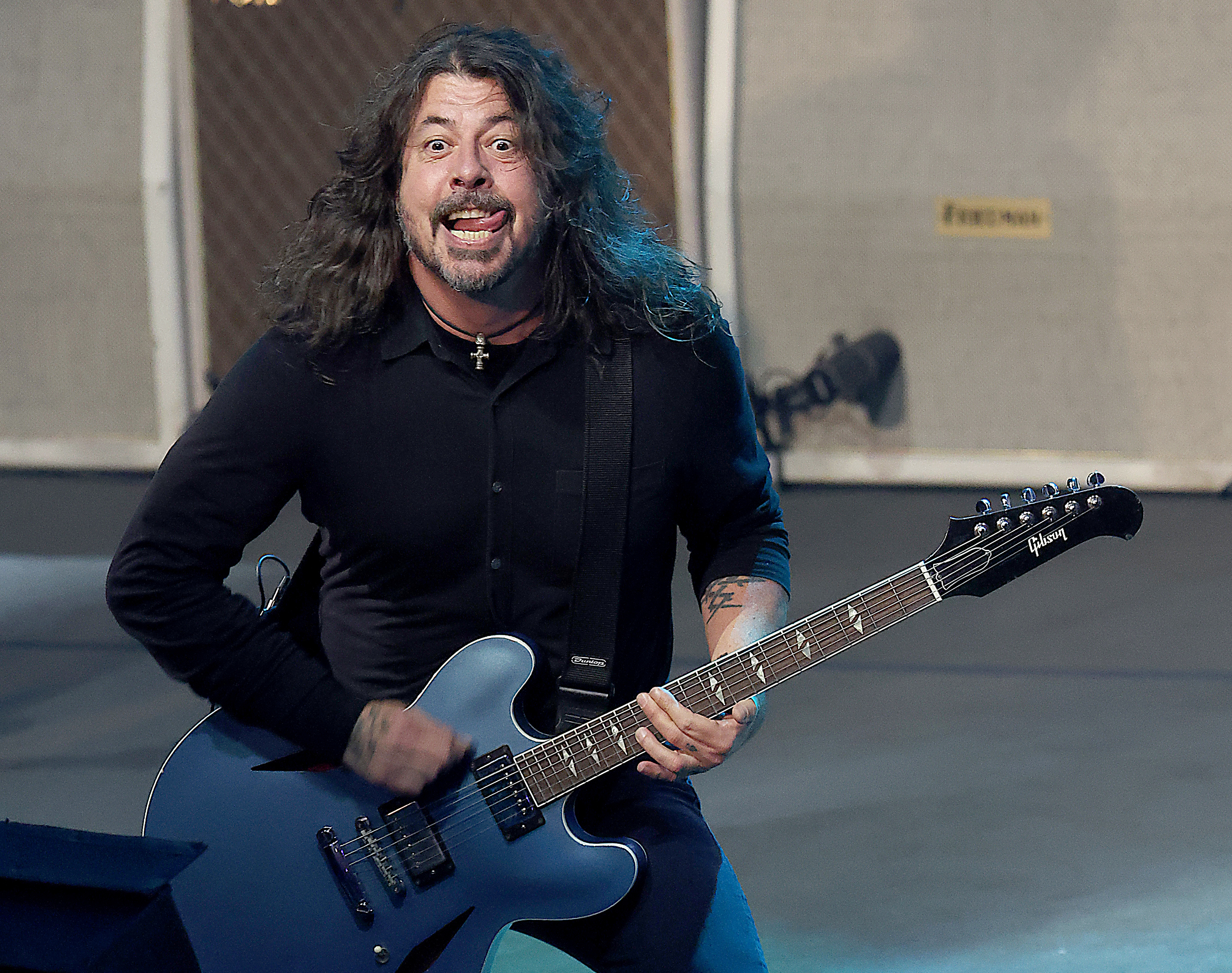 Dave Grohl energetically playing an electric guitar on stage. He is smiling widely, wearing a long-sleeve shirt, and has long wavy hair