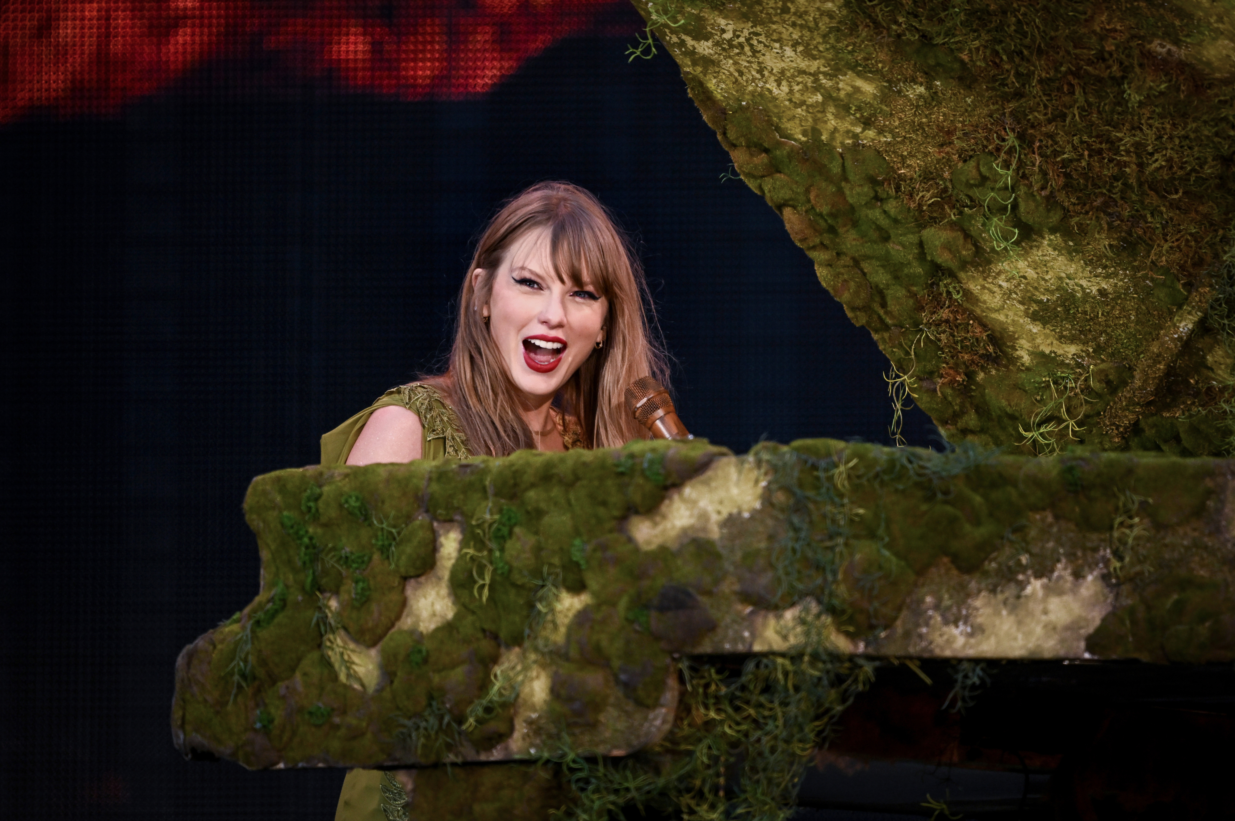 Taylor Swift performs while playing a piano covered in moss
