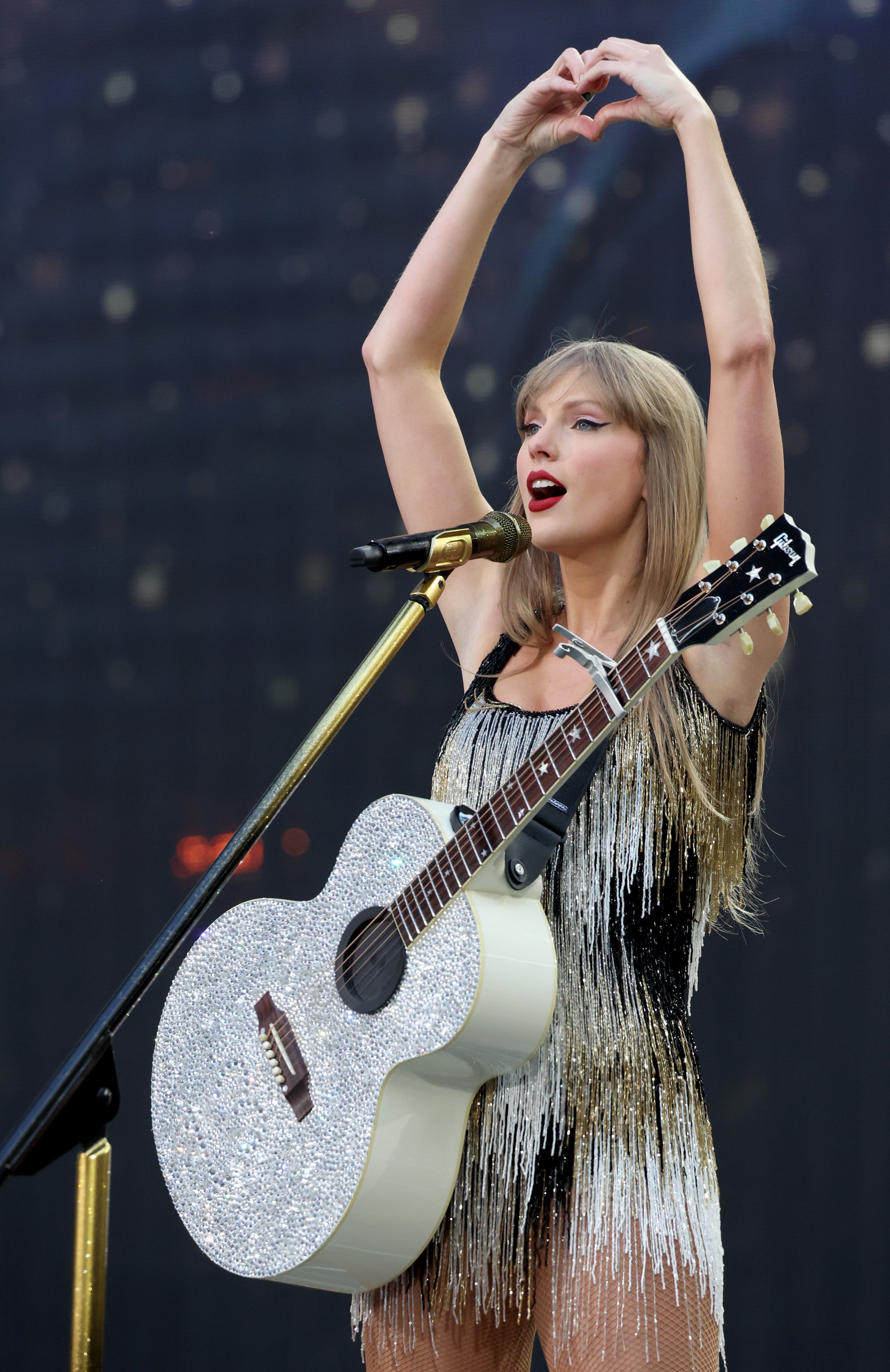 Taylor Swift on stage, wearing a fringed outfit, holding a glittery guitar, making a heart shape with her hands above her head