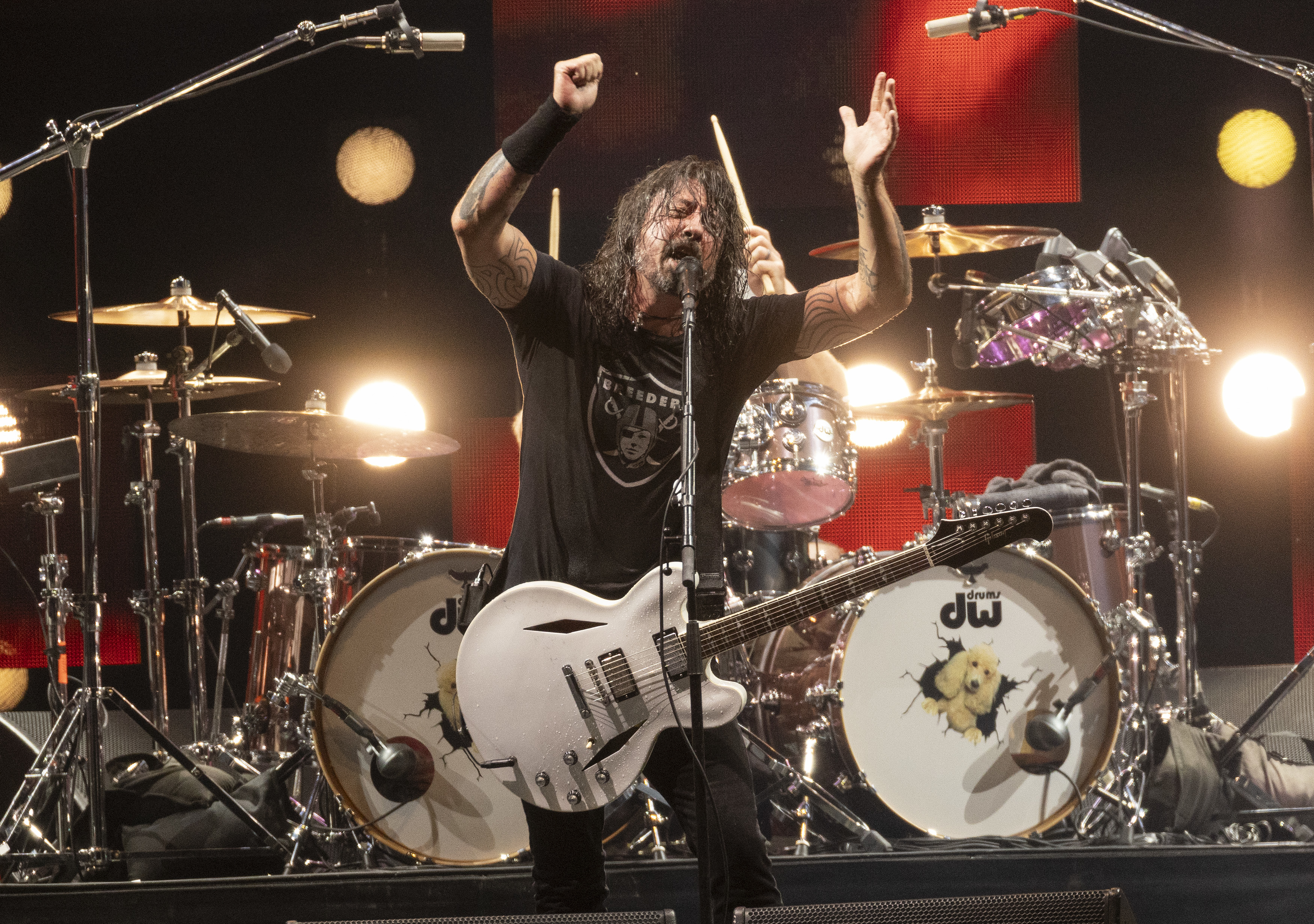 Dave Grohl performing on stage with a white electric guitar, drum set and cymbals in the background