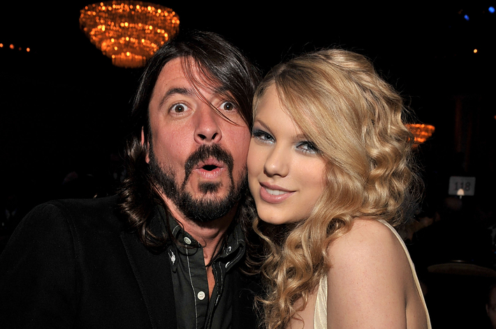 Taylor Swift in a sleeveless dress posing with Dave Grohl in a suit at an event. Both are smiling