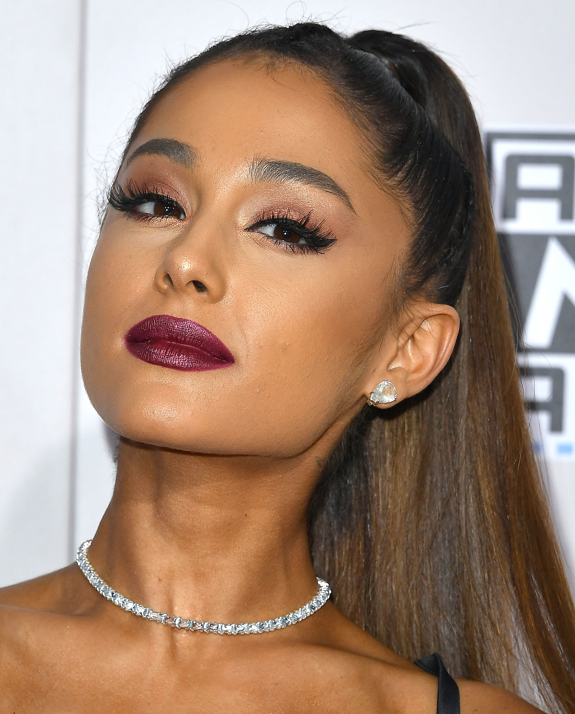 Ariana Grande, wearing a diamond choker necklace and stud earrings, poses with her hair in a high ponytail and bold makeup on the red carpet