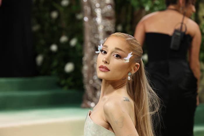 Ariana Grande poses on a green carpet, wearing a strapless dress and dramatic white feathered eyelashes, with her hair styled long and straight