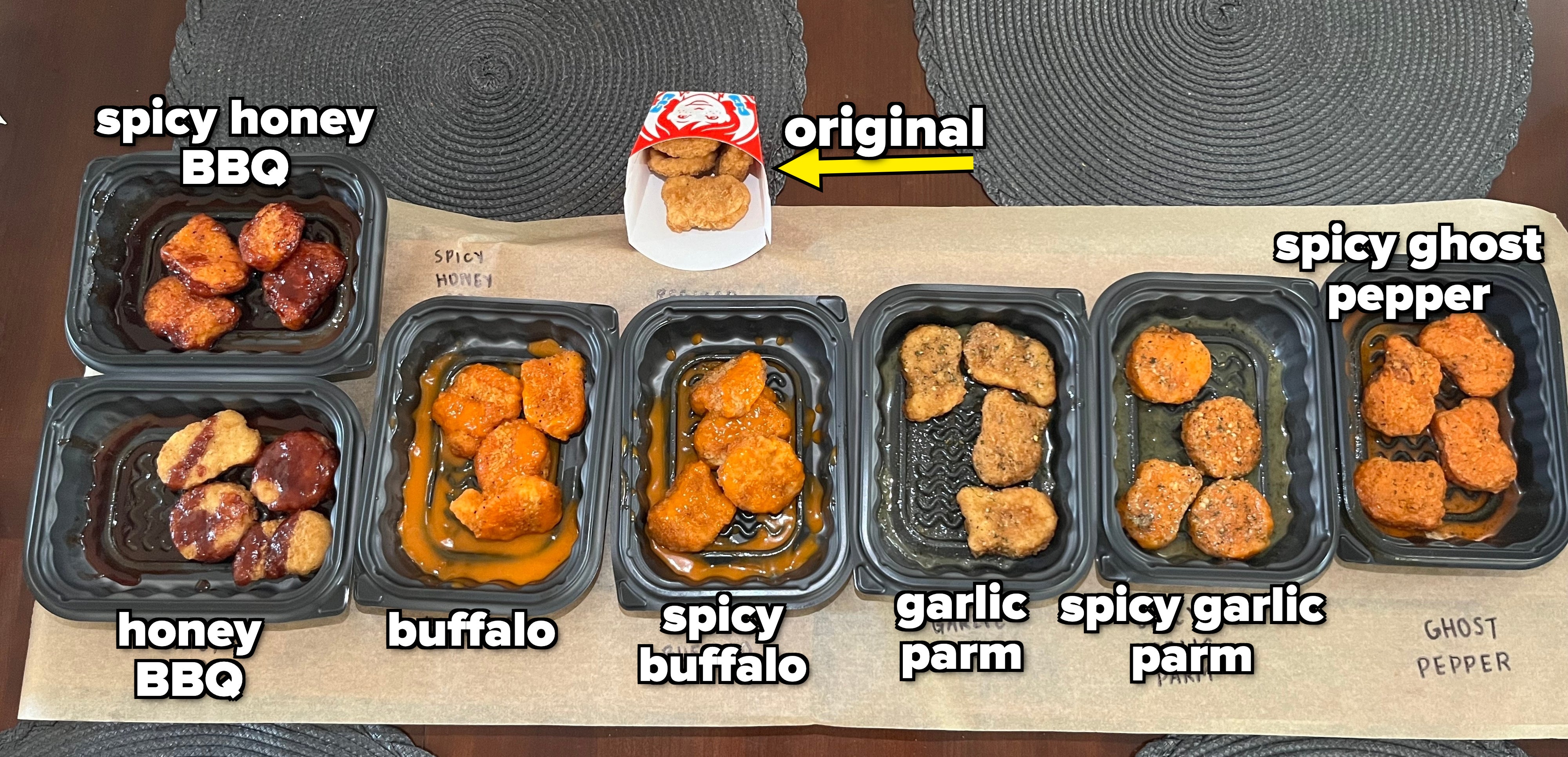 Seven containers of chicken wings labeled Honey BBQ, Spicy Korean, Buffalo, Buffalo (another type), Garlic Parm, Spicy Garlic Parm, and Ghost Pepper are displayed on a table