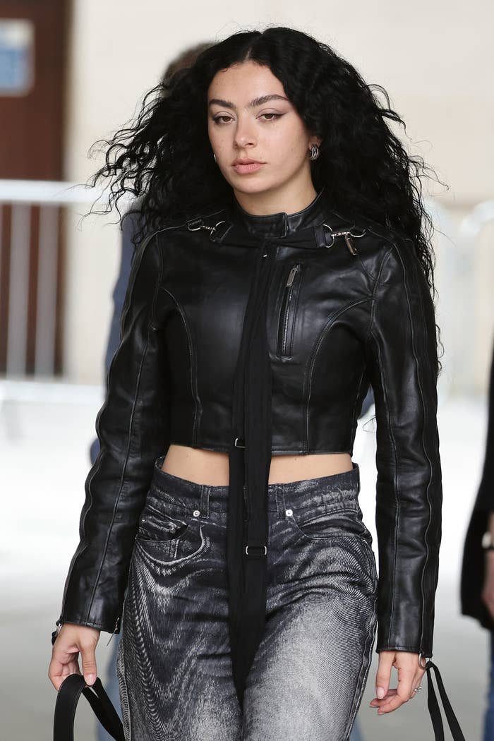 Charli XCX is wearing a black leather cropped jacket and high-waisted jeans. She is walking outdoors