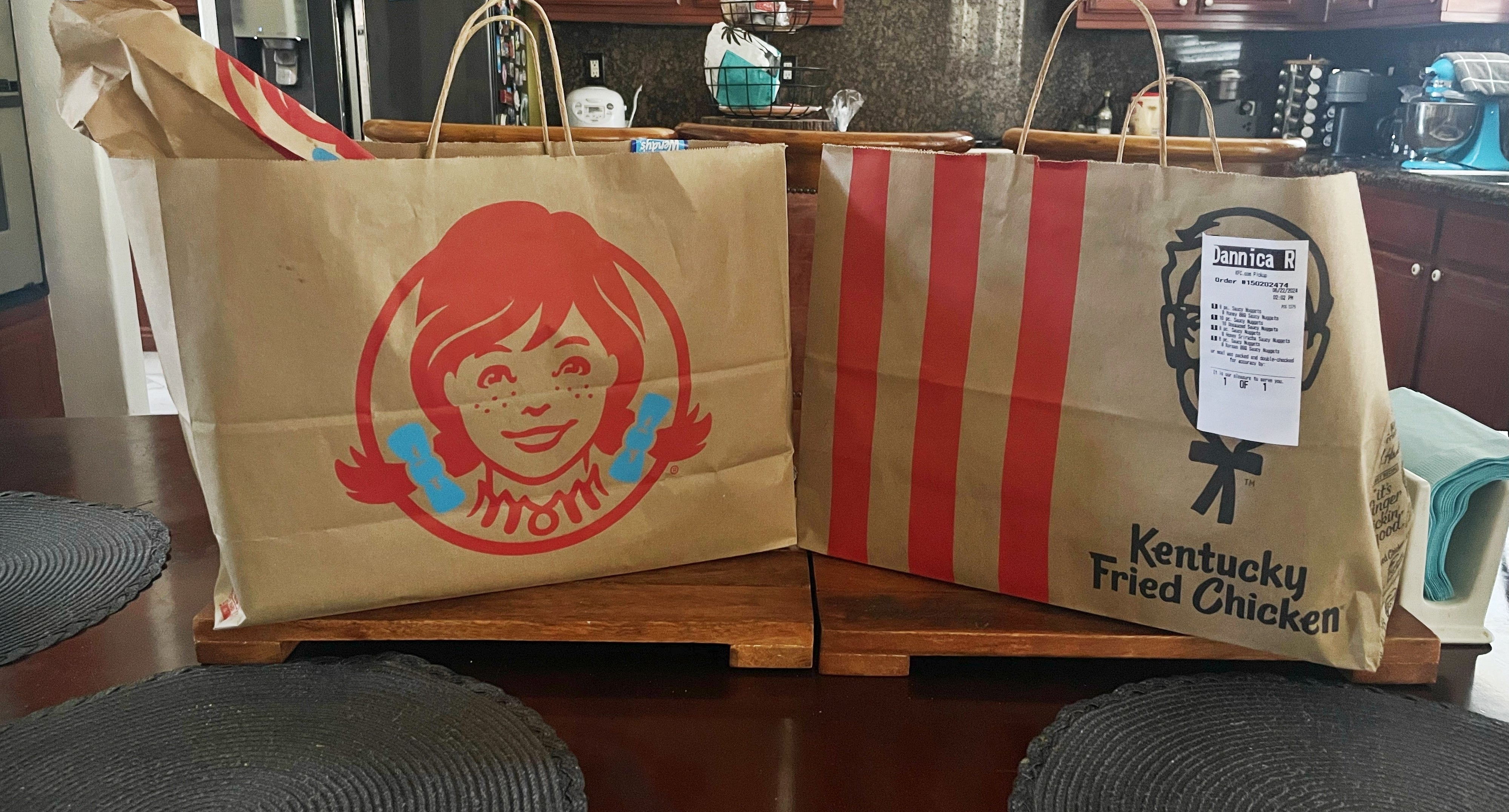 Two large takeout bags, one from Wendy&#x27;s and one from Kentucky Fried Chicken, are placed on a kitchen island with wooden cabinets and a refrigerator in the background