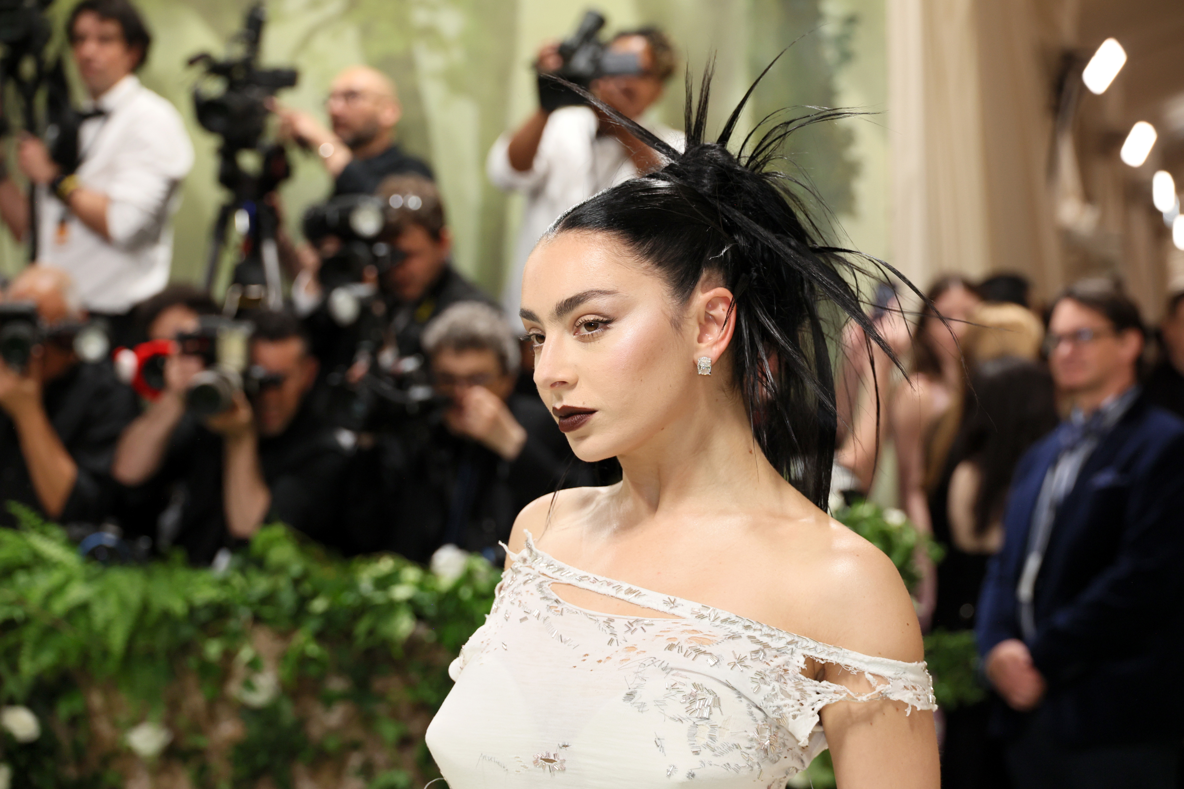 Charli XCX on the red carpet, wearing a white lace dress with off-the-shoulder design and elaborate black hairpiece, surrounded by photographers
