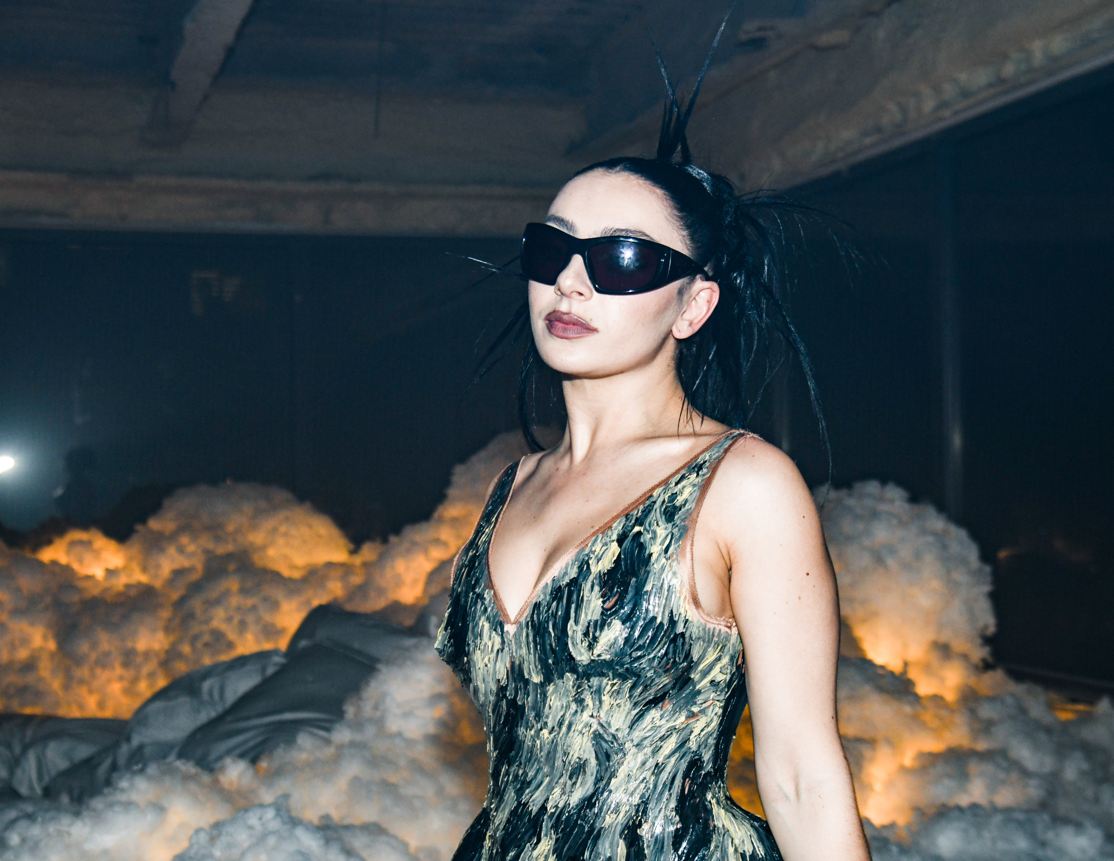 Charli XCX wearing a modern, artistic dress with black sunglasses and spiky hair, posing indoors with a cloud-like backdrop