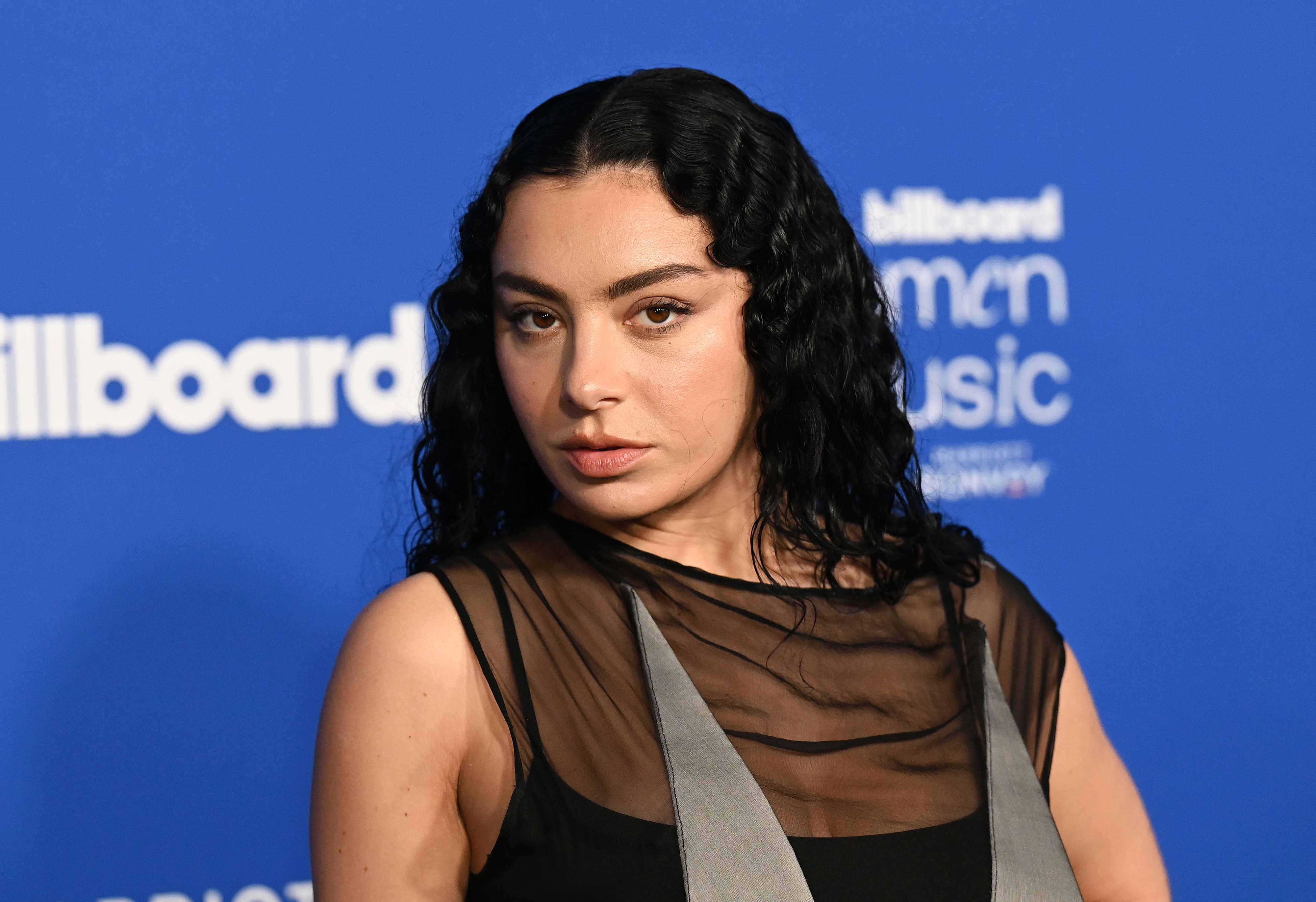 Charli XCX at Billboard Women in Music event, wearing a black dress with a sheer top and gray accents, standing against a blue background with Billboard logos