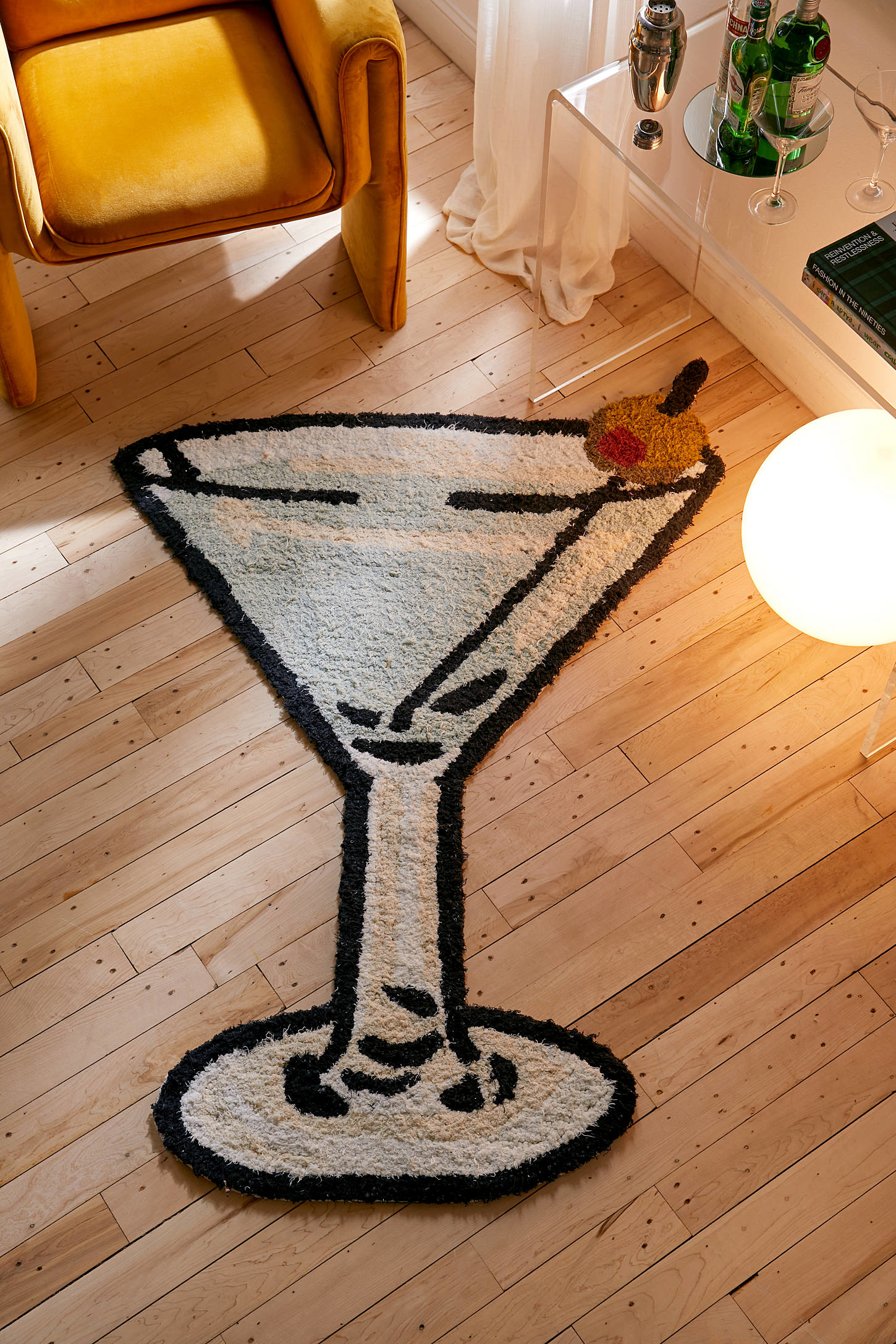 Martini-themed rug shaped like a cocktail glass with an olive, placed next to a chair, lamp, and table with drinks. Ideal for stylish home decor