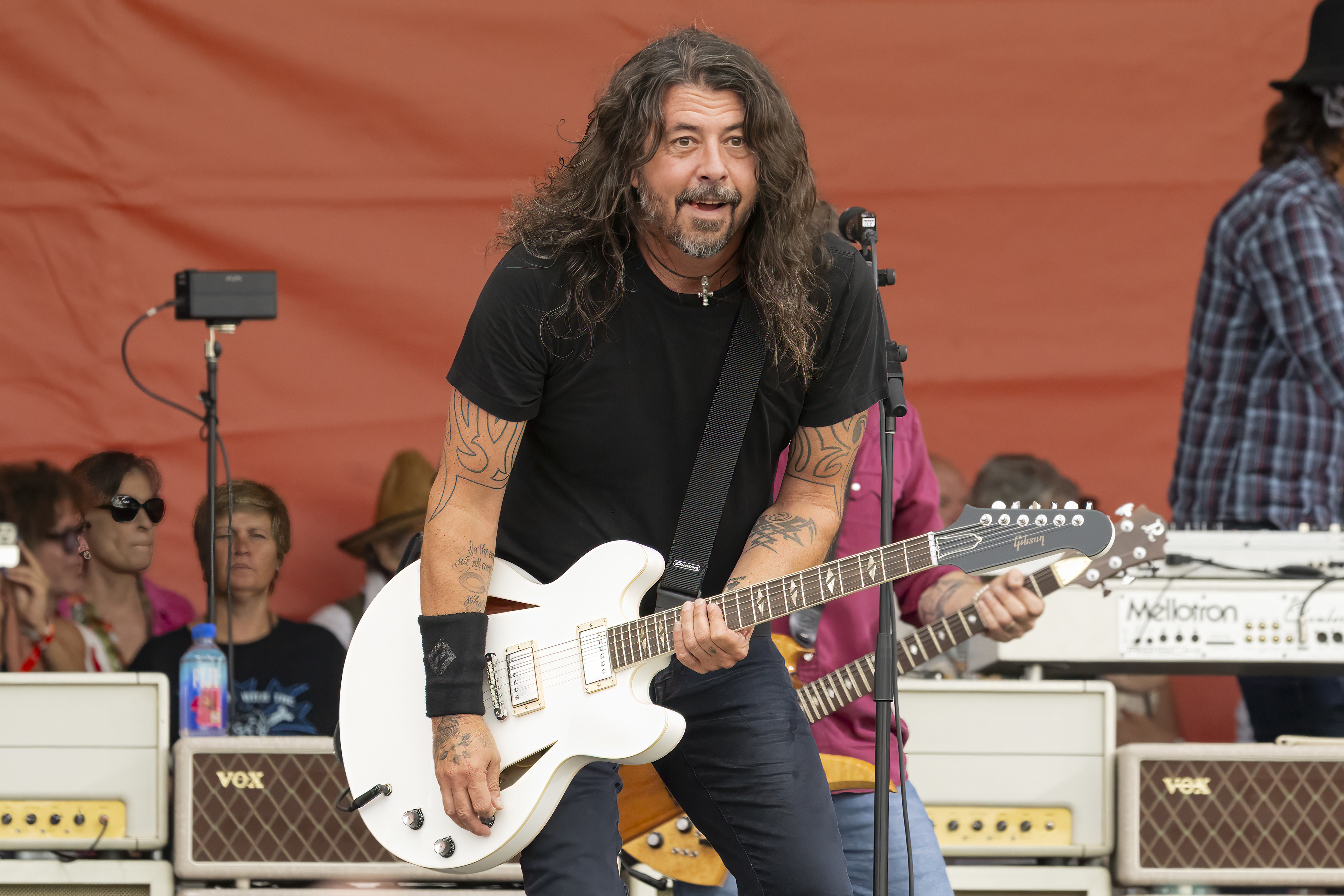 Dave Grohl on stage performing with a white electric guitar. Other band members and equipment visible in the background