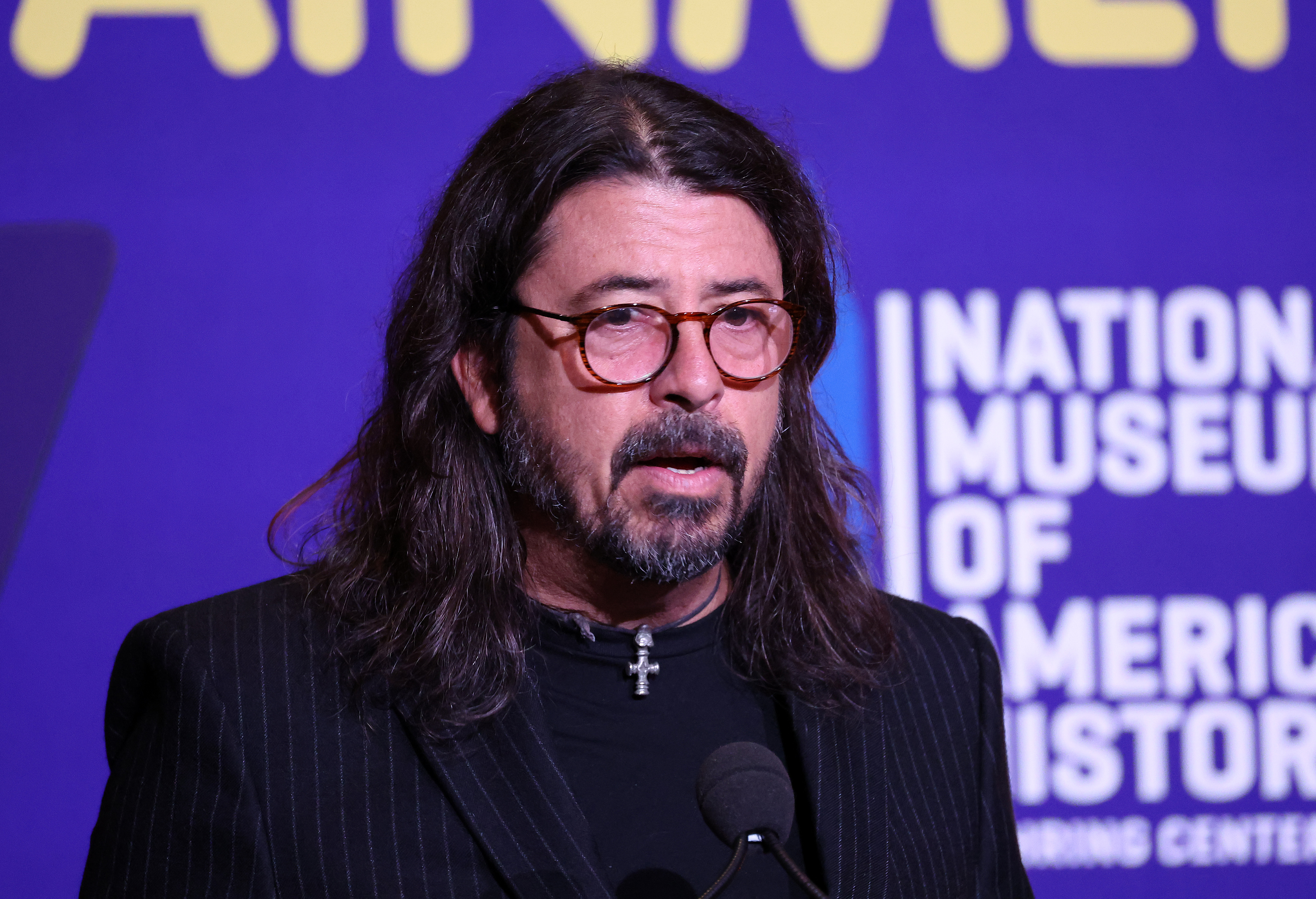 Dave Grohl speaks at an event in front of a sign for the National Museum of American History, wearing glasses and a black pinstriped suit jacket