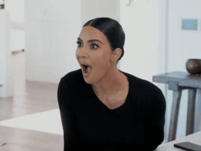 Kim Kardashian sits indoors wearing a black top, looking surprised with her mouth open