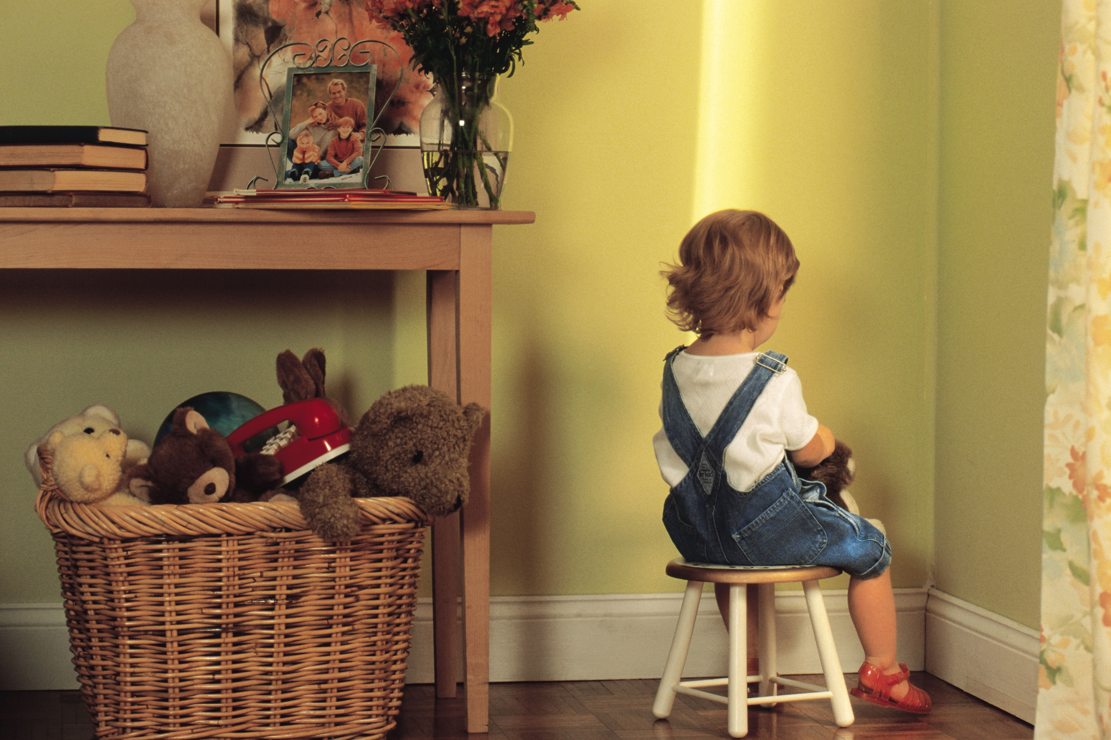 A young child sits on a stool facing the corner of a room. A basket of stuffed animals and toys is on the floor nearby