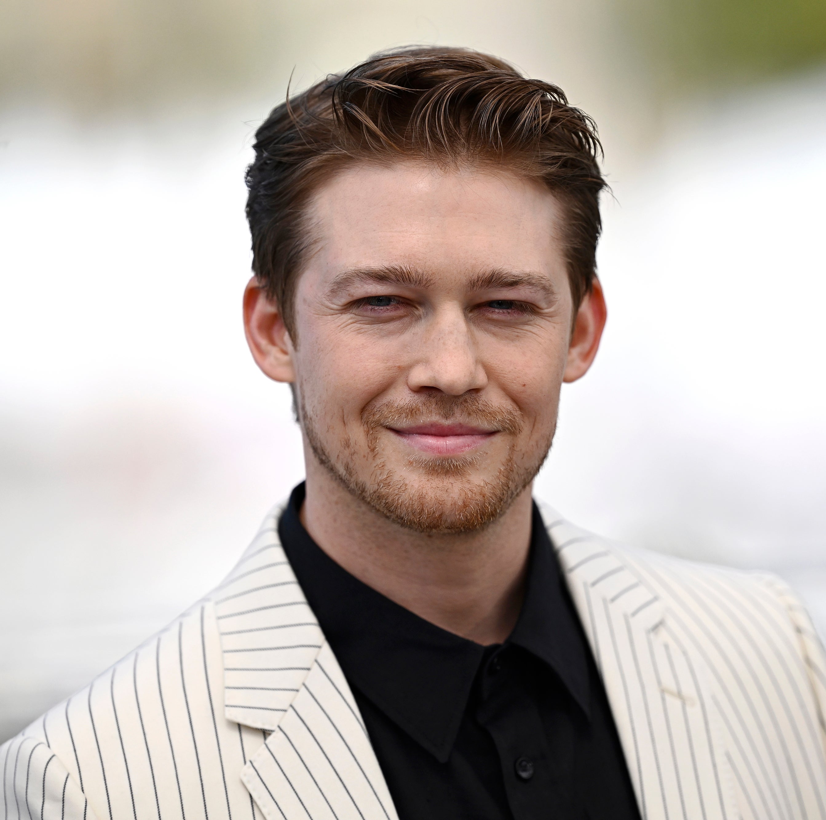 Joe Alwyn is pictured outdoors, wearing a light-colored, pinstriped jacket and a dark shirt, smiling gently with a blurred background