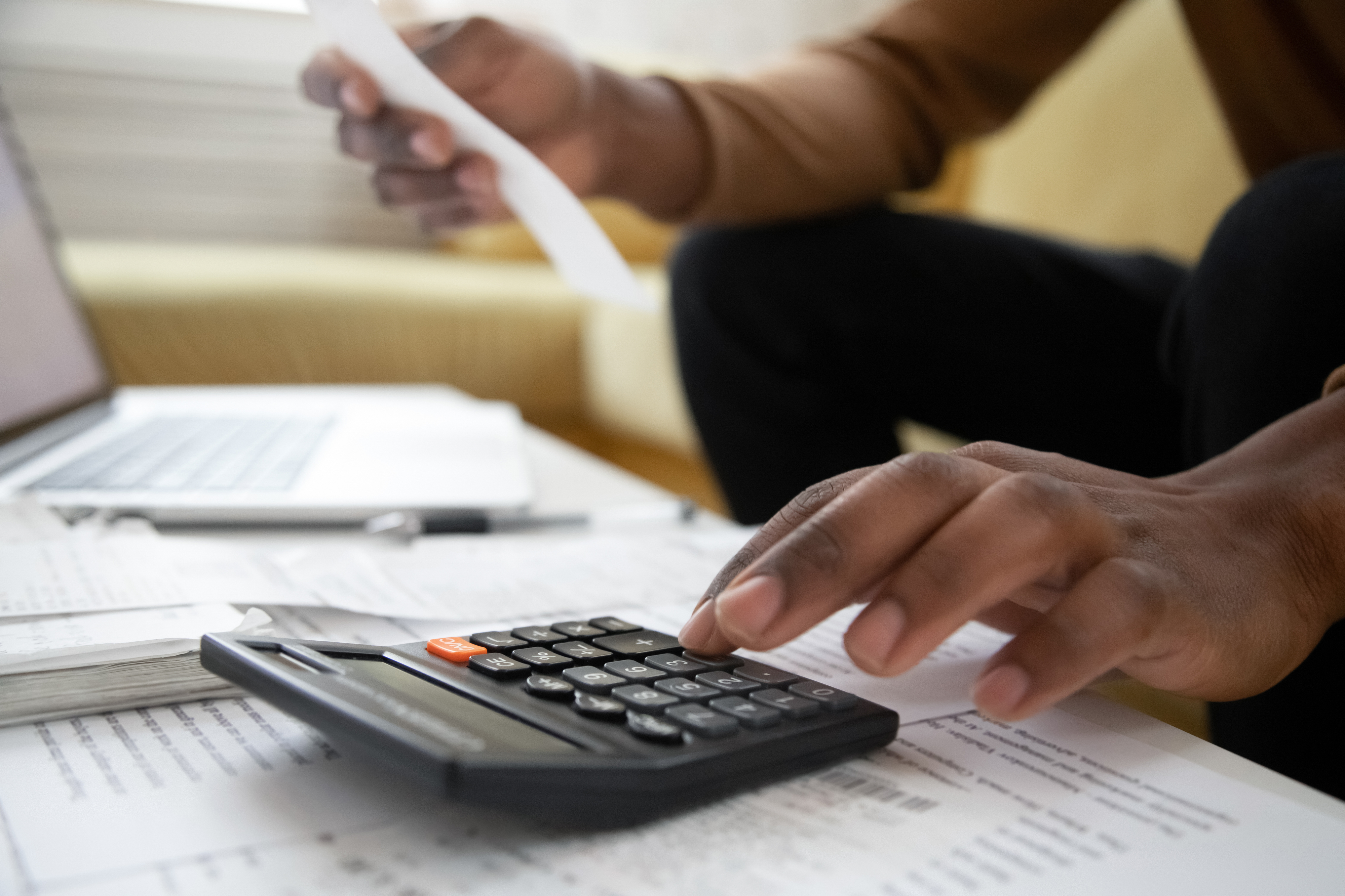 A person calculating finances with a calculator and holding a receipt, surrounded by papers and a laptop on a table