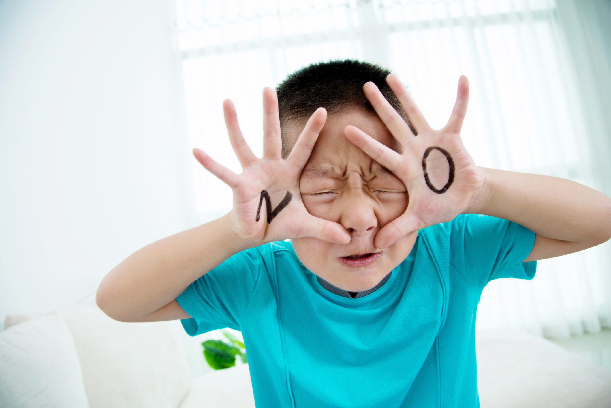 Child in a blue shirt holding their hands in front of their face with &quot;N&quot; and &quot;O&quot; written on their palms, expressing a playful refusal