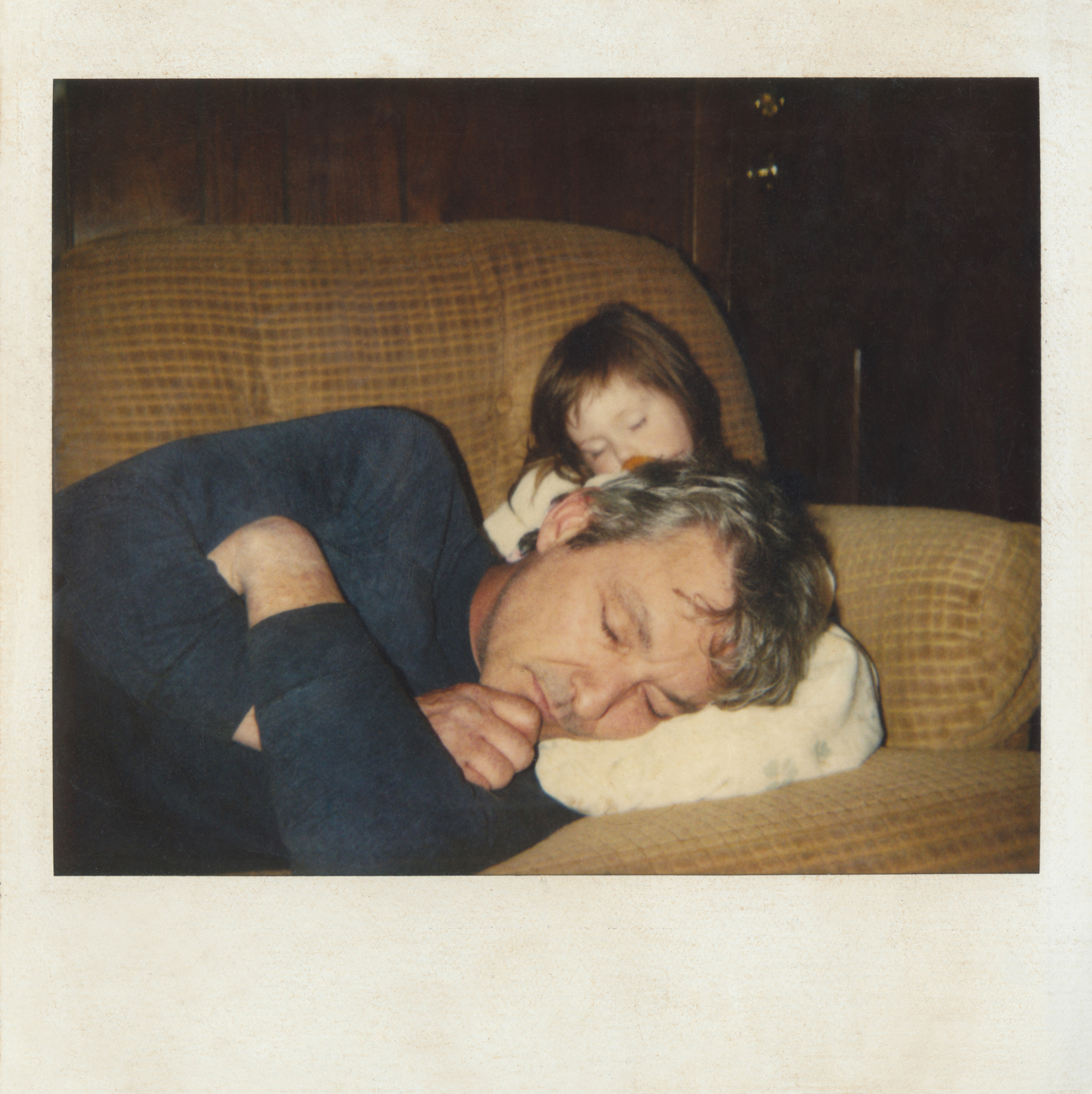 Man and young child sleeping on a couch, with the man&#x27;s head resting on a cushion and the child resting on his shoulder