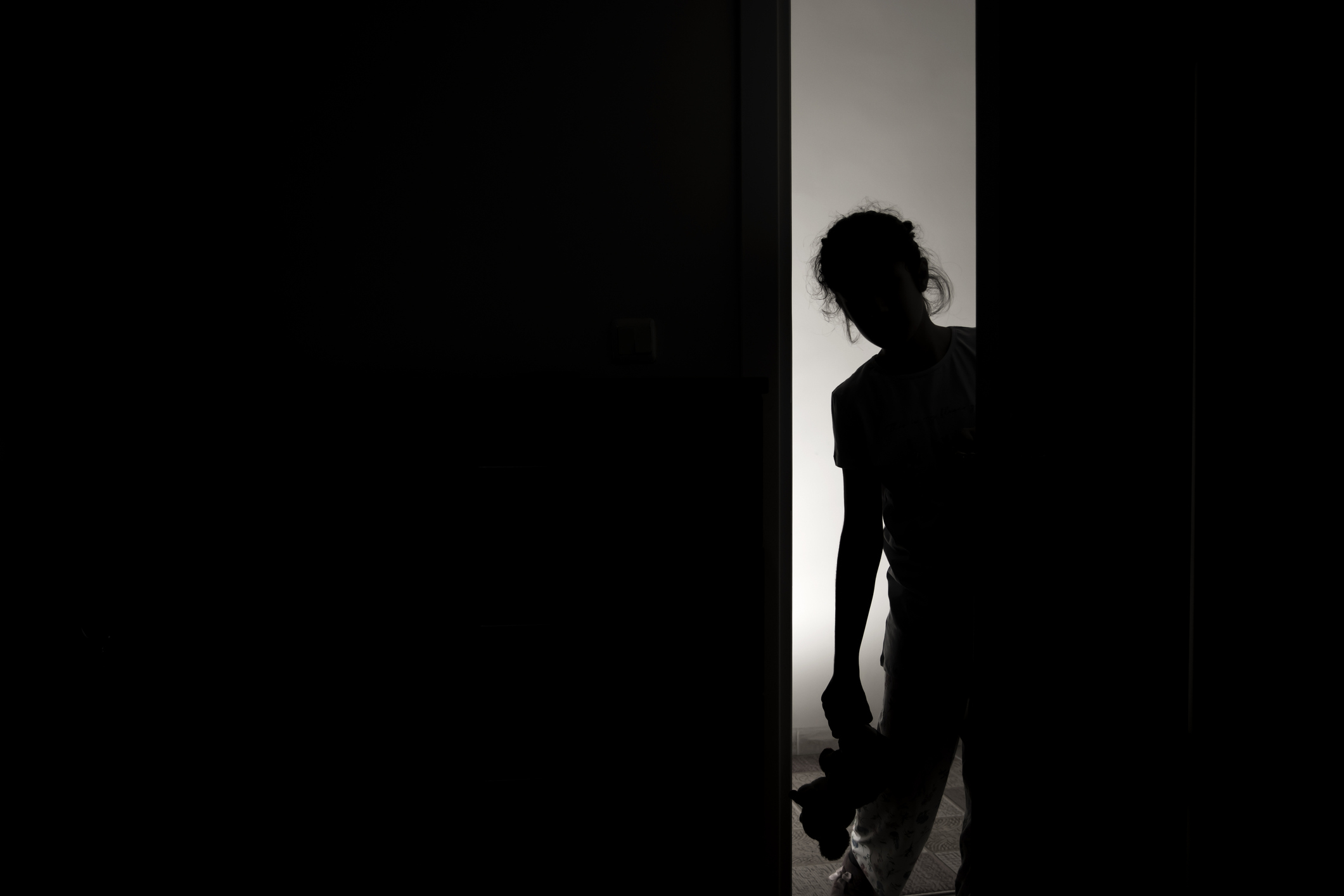 Silhouette of a child holding a teddy bear, standing in a doorway partially illuminated from behind