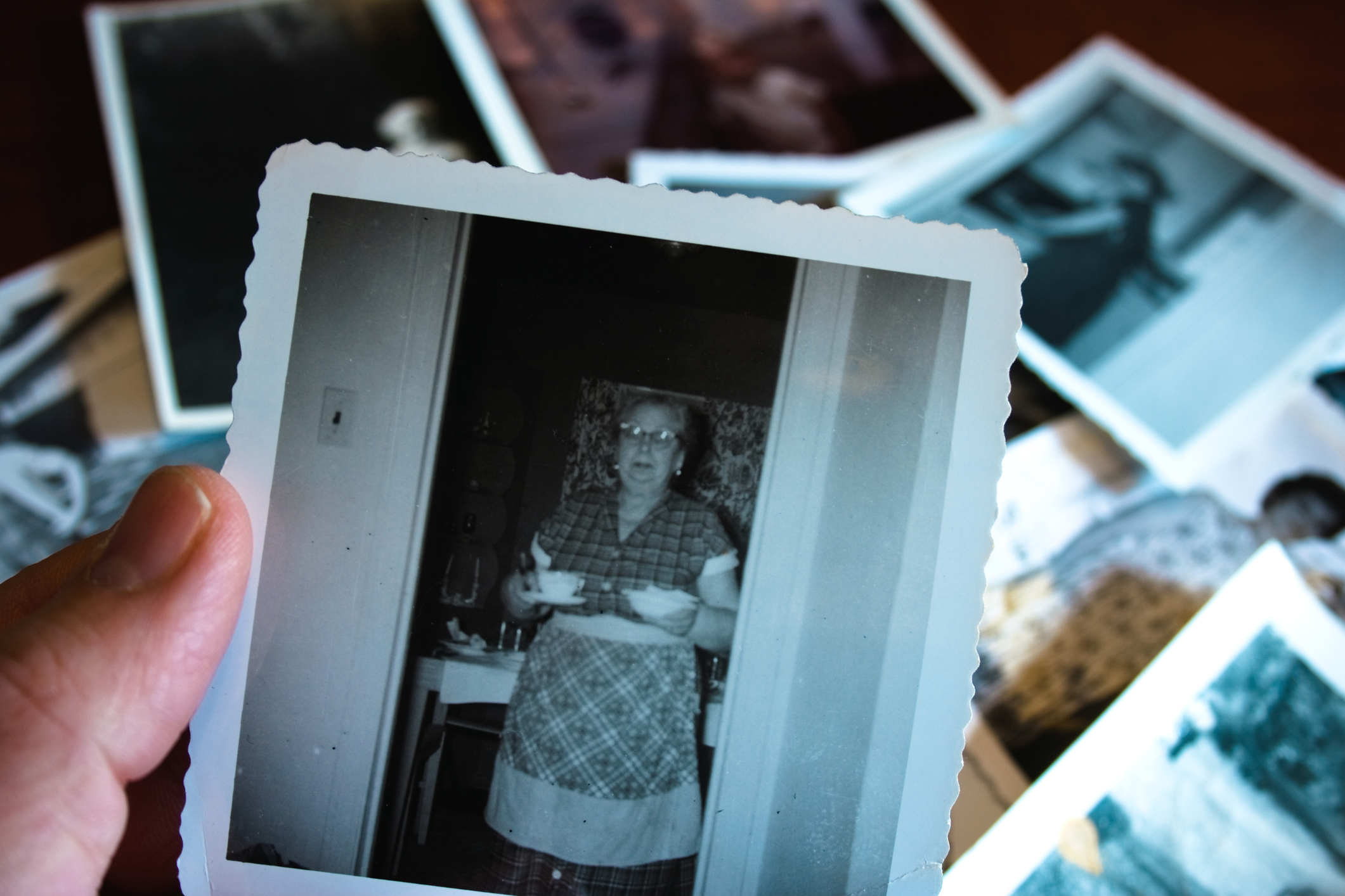 A hand holds a vintage black-and-white photograph of an elderly woman in a patterned dress, standing in a doorway. The background shows more scattered photographs