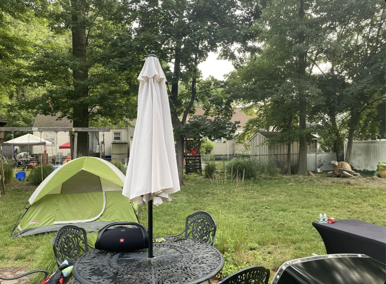 Outdoor backyard scene with a green tent, patio table with an umbrella, and an enclosed garden area. Trees and a house are visible in the background