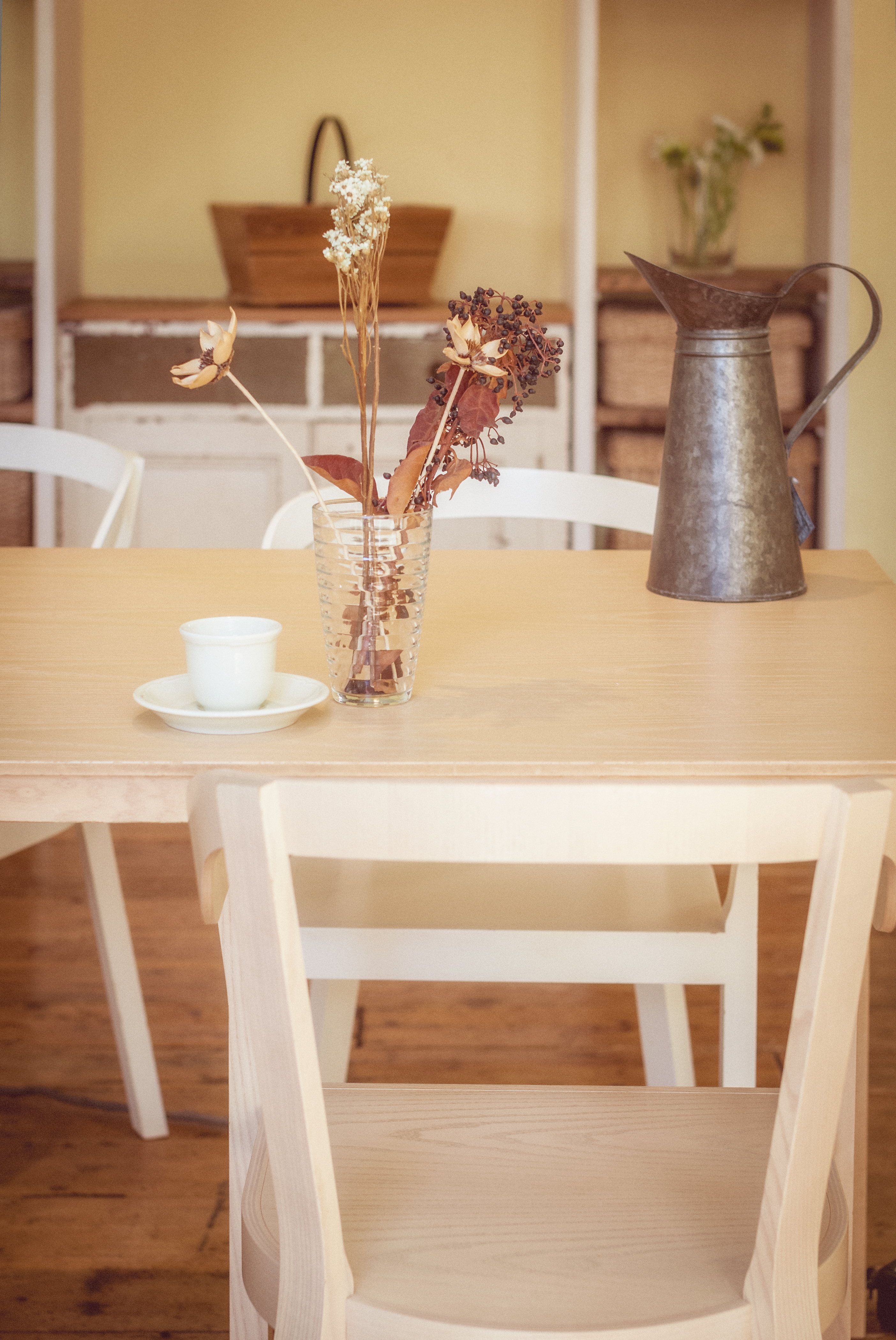 Minimalist dining table with dried flowers in a vase, a white teacup on a saucer, and a metal pitcher in the background